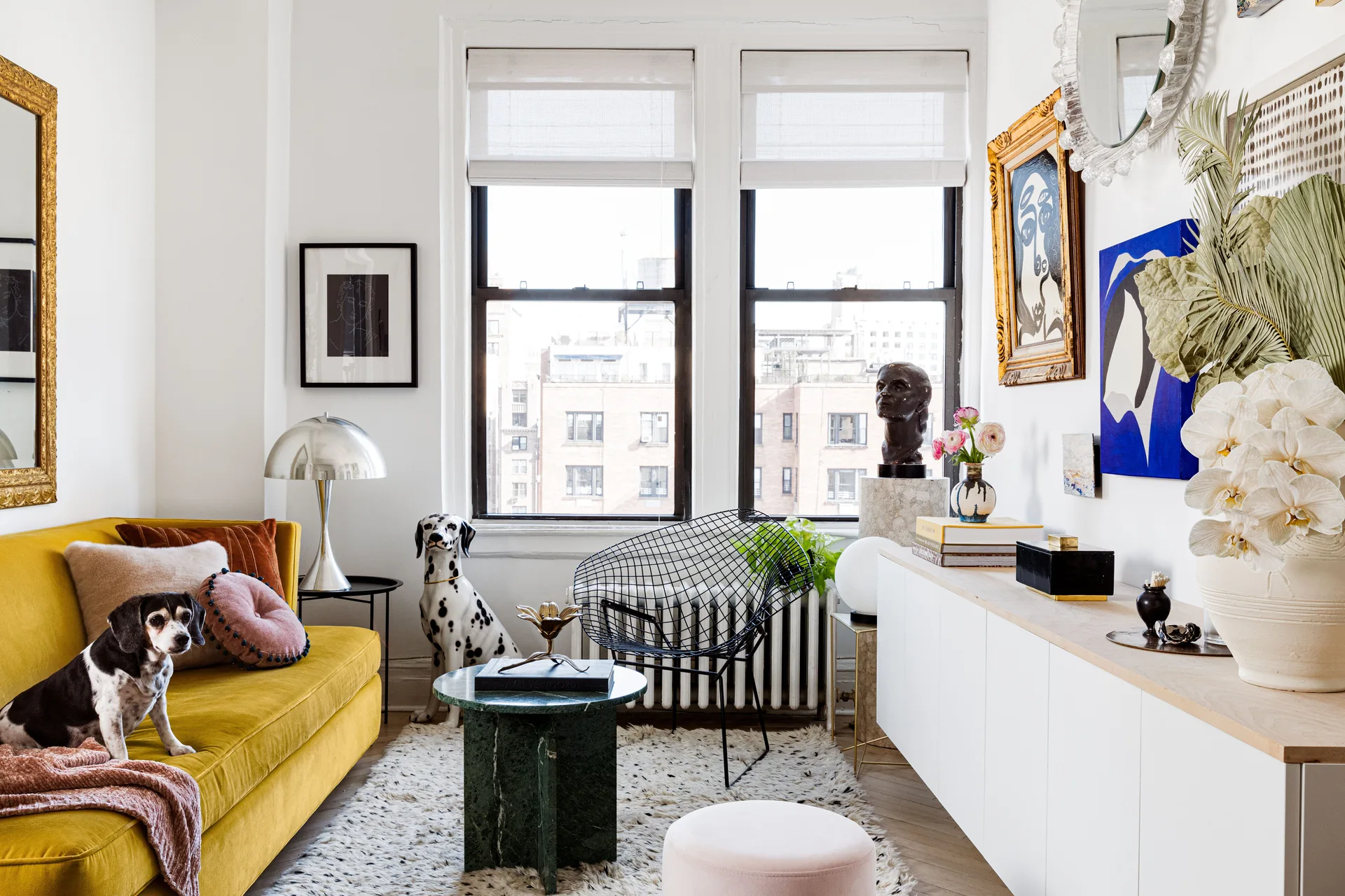 A bright living room features a yellow velvet sofa, a green marble coffee table, and a white console under a display of eclectic artwork.