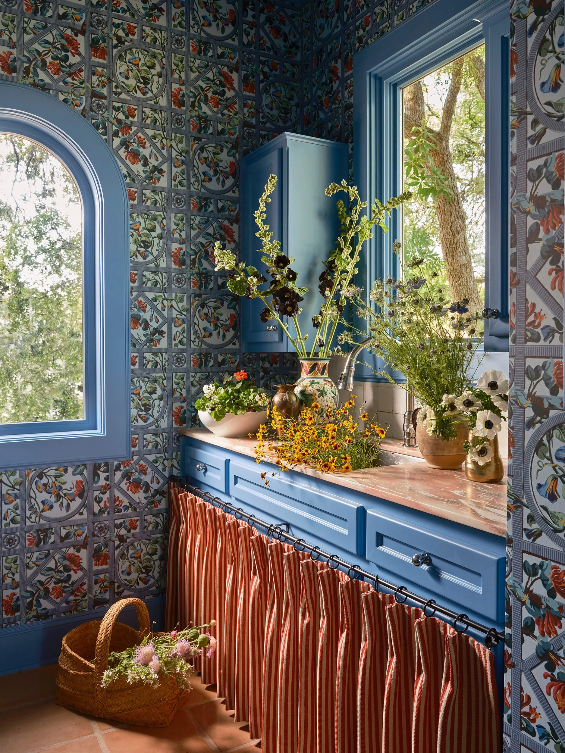 A utility room features blue cabinetry, colorful floral wallpaper, an under-sink striped fabric skirt, and terracotta floor tiles.