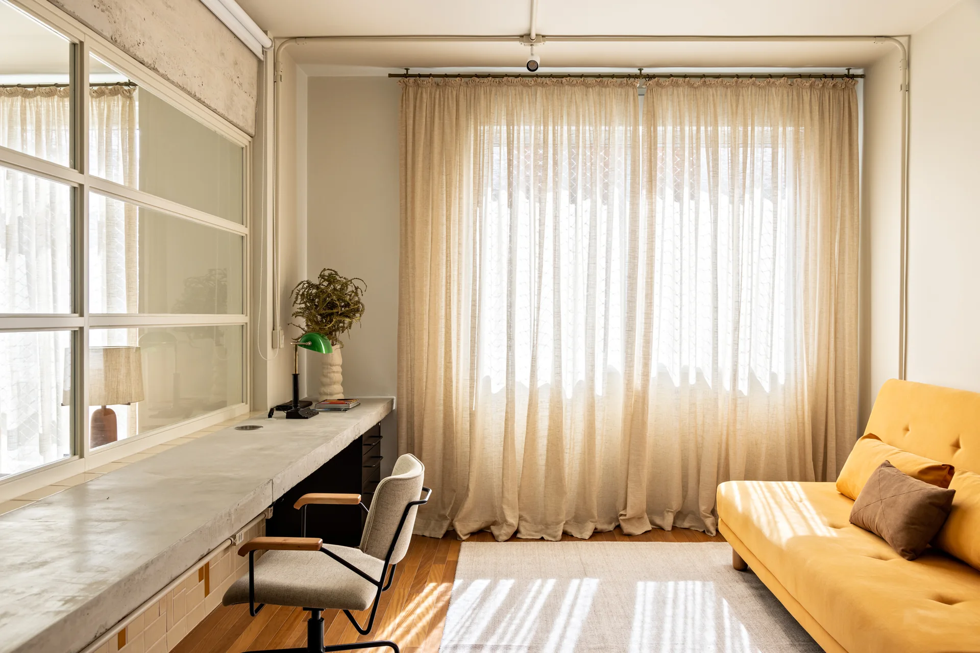 A minimalist multi-purpose room features a long concrete desk, an upholstered office chair, and a bright yellow sofa bed beneath sheer curtains.