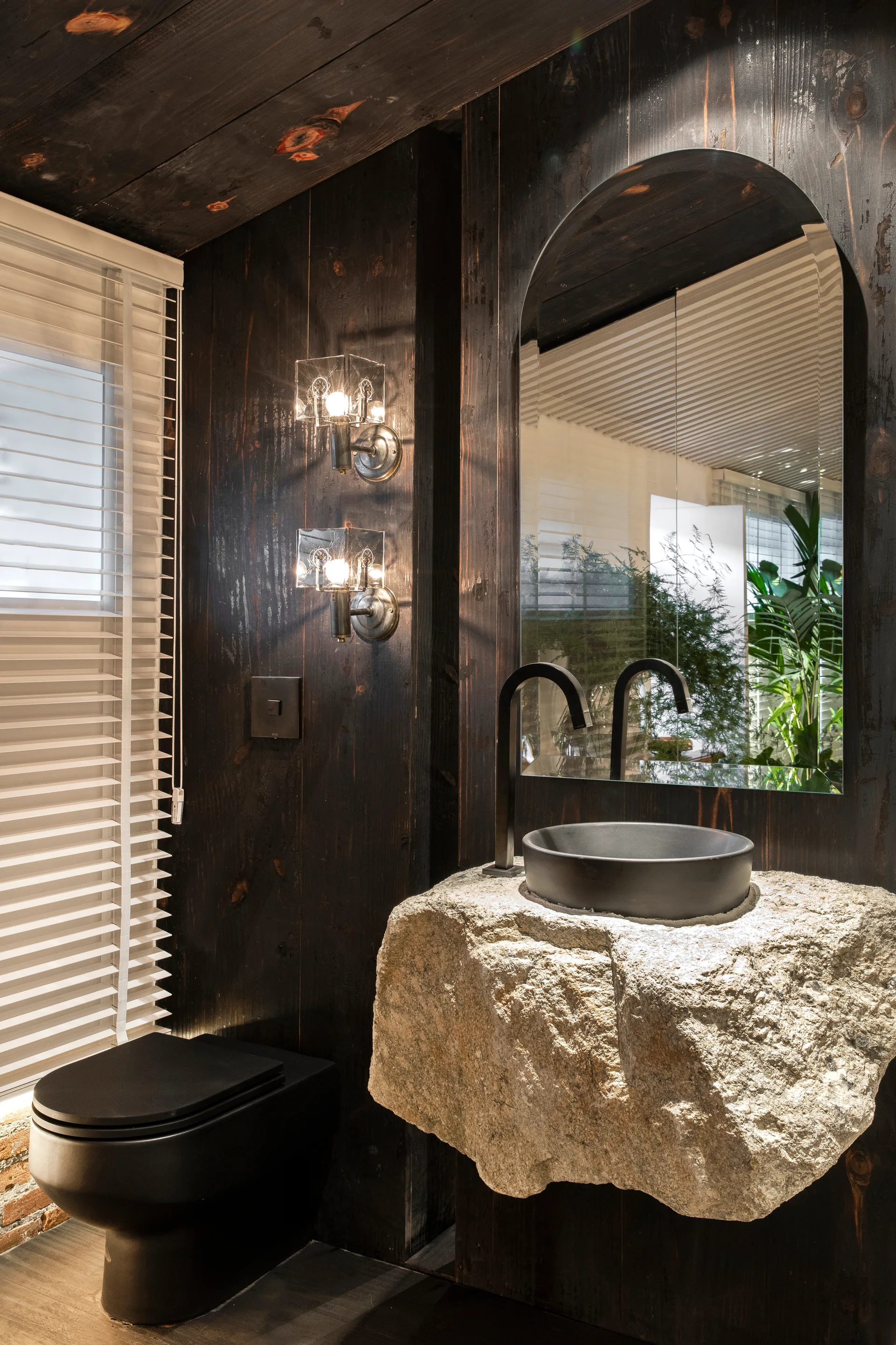A powder room features a rough-hewn stone vanity with a black basin and faucet, dark wood-paneled walls, an arched mirror, and a matte black toilet.