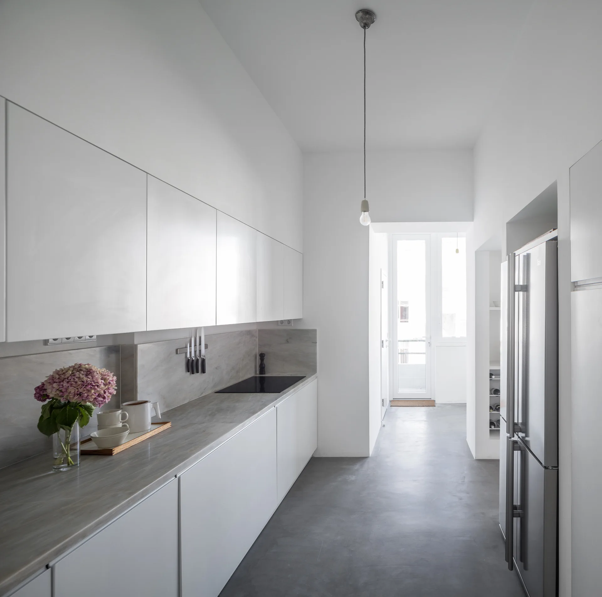 A minimalist galley kitchen featuring white handleless cabinets, a grey stone countertop and backsplash, and smooth grey concrete flooring.