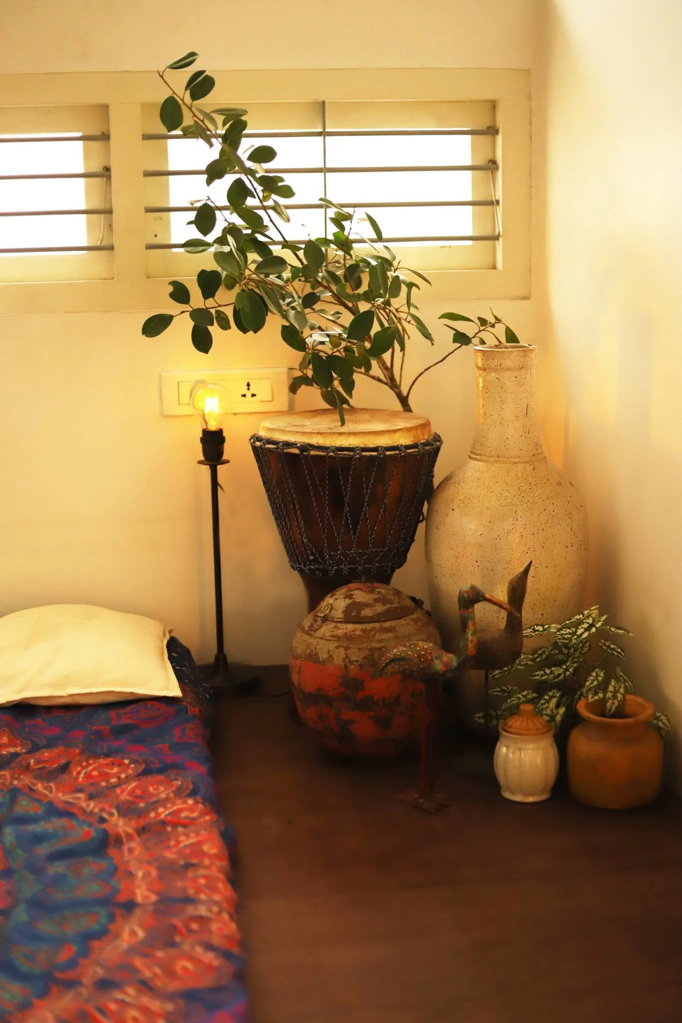 A cozy corner vignette features a djembe drum, a tall speckled ceramic vase, potted plants, and an exposed bulb lamp next to a floor cushion.
