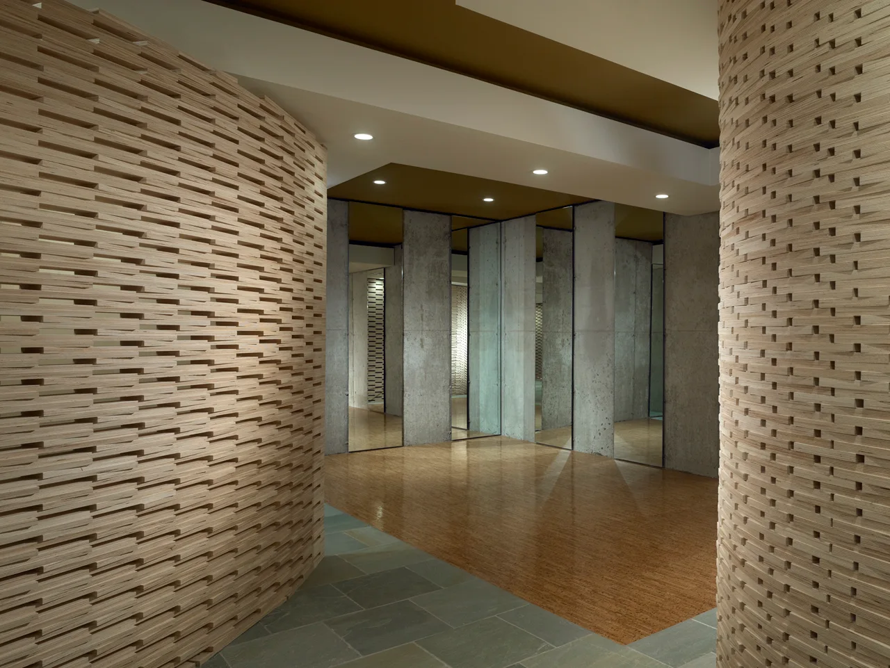 A modern hallway features curved walls made of stacked horizontal plywood strips, mirrored columns, and a floor transition from slate to warm wood.