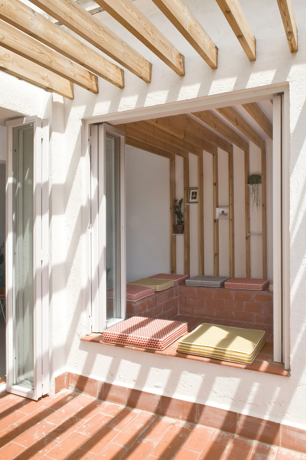 An outdoor patio features a built-in red brick bench with patterned cushions under a wooden pergola casting shadows on terracotta tile flooring.