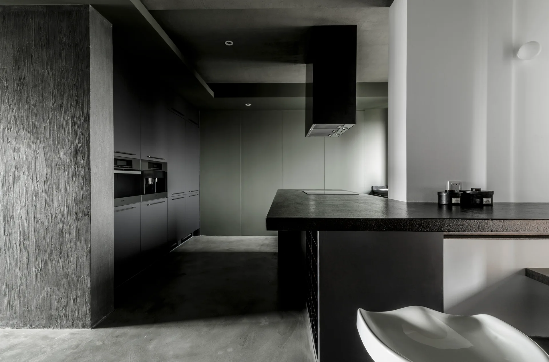 A minimalist modern kitchen with matte black cabinetry, a dark textured pillar, concrete floors, and a black kitchen island with a range hood.