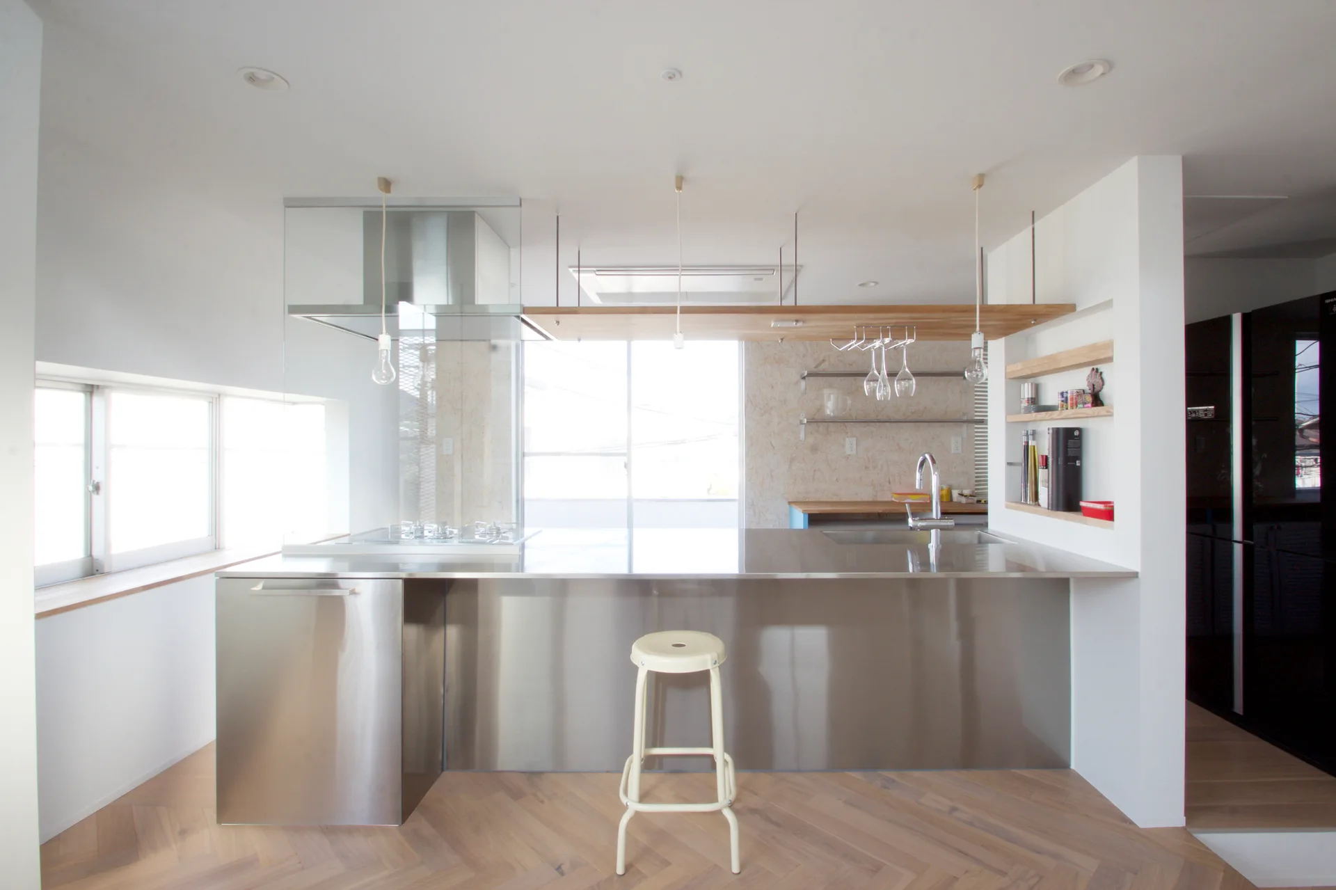 A minimalist white kitchen featuring a large stainless steel island with a single stool and light herringbone wood floors.