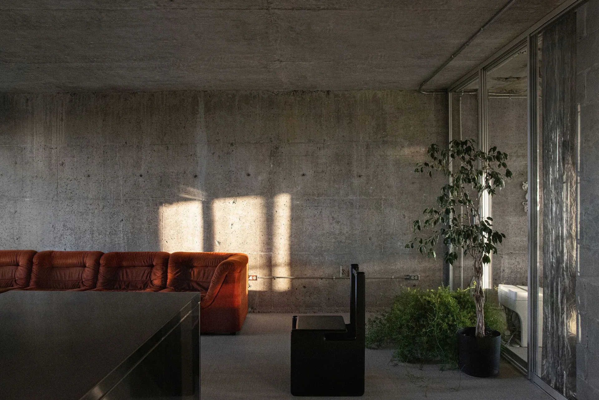 A minimalist living room featuring a long red sofa, a black chair, and potted plants set against raw concrete walls and ceiling.