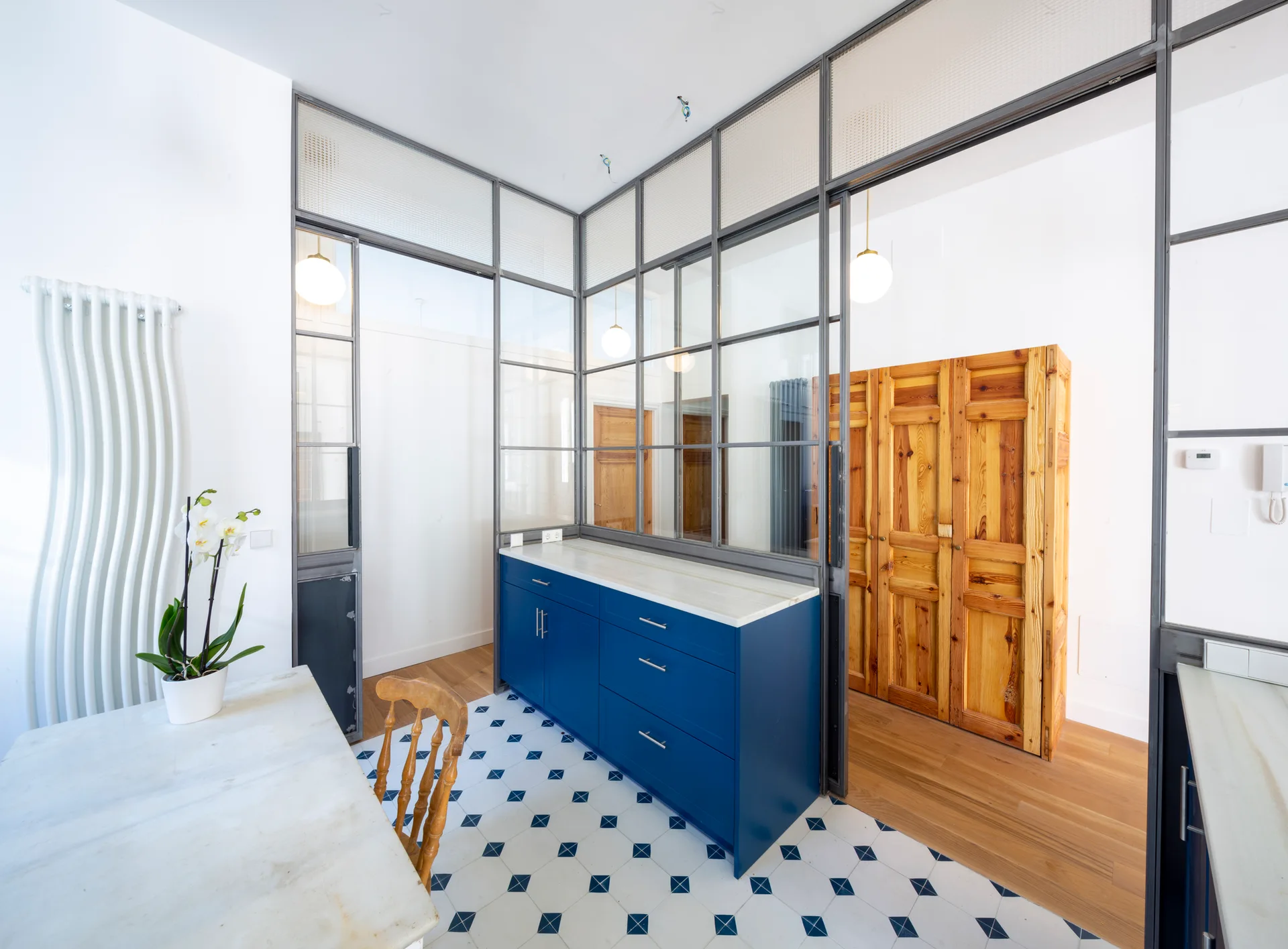 A kitchen area featuring blue cabinets, a geometric tiled floor, and a metal-framed glass partition separating it from a wood-floored space.
