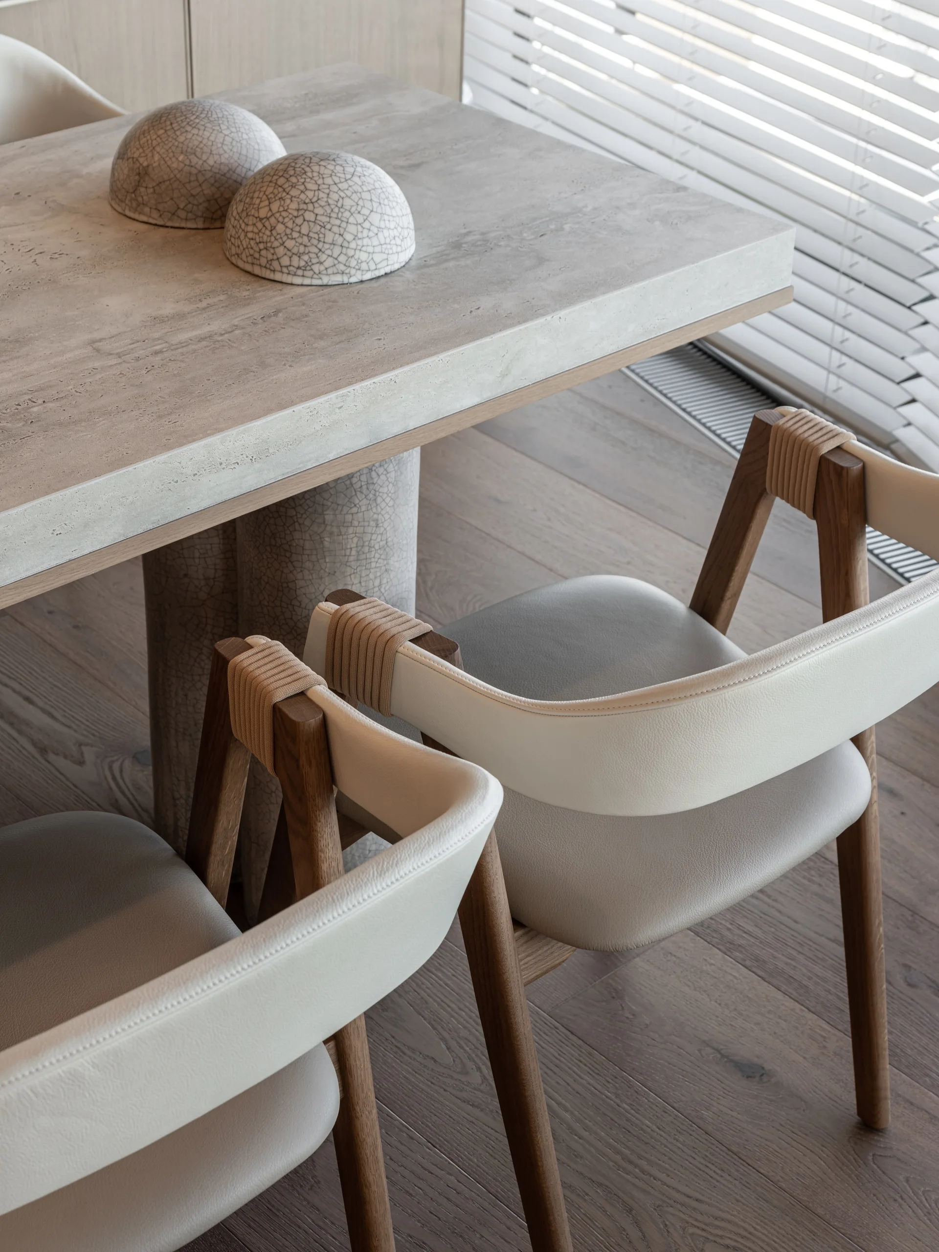 Modern dining area featuring a stone-topped table, decorative textured bowls, and wood frame chairs with leather backrests and woven cord accents.
