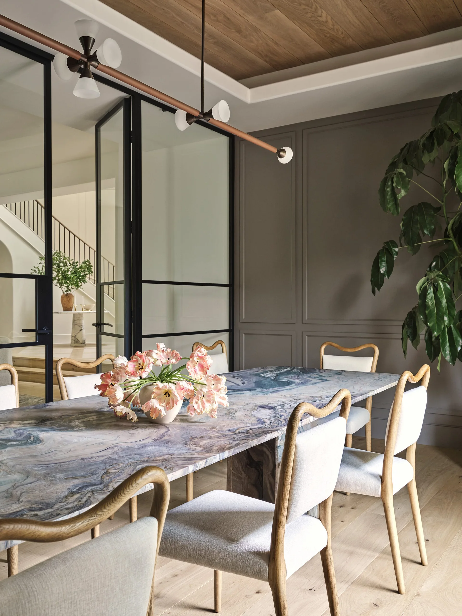 A dining room featuring a blue swirled marble table, light wood chairs, paneled grey walls, and black-framed glass partition doors.
