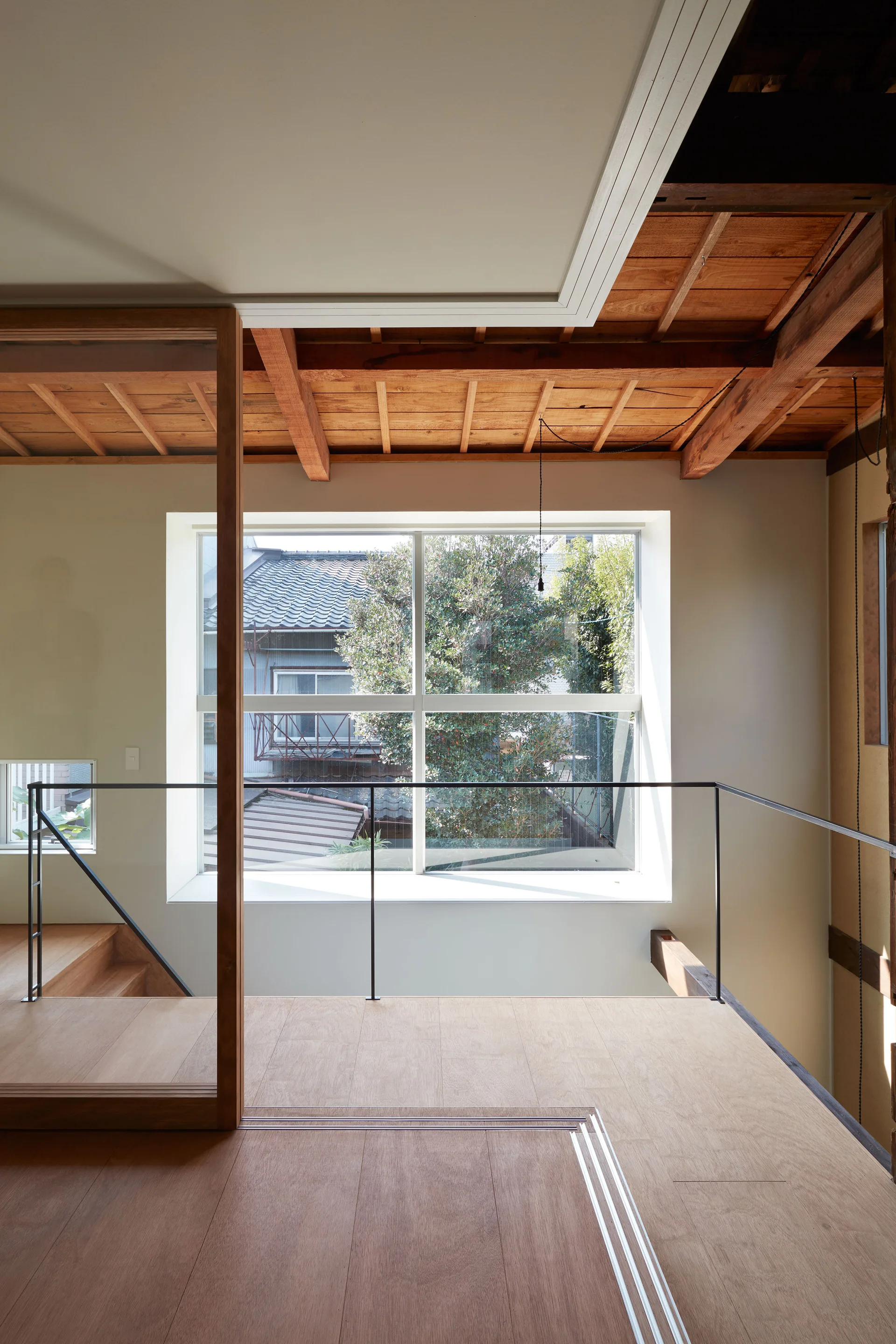A minimalist home landing features light wood flooring, a thin black metal railing, and a large window beneath an exposed timber ceiling.