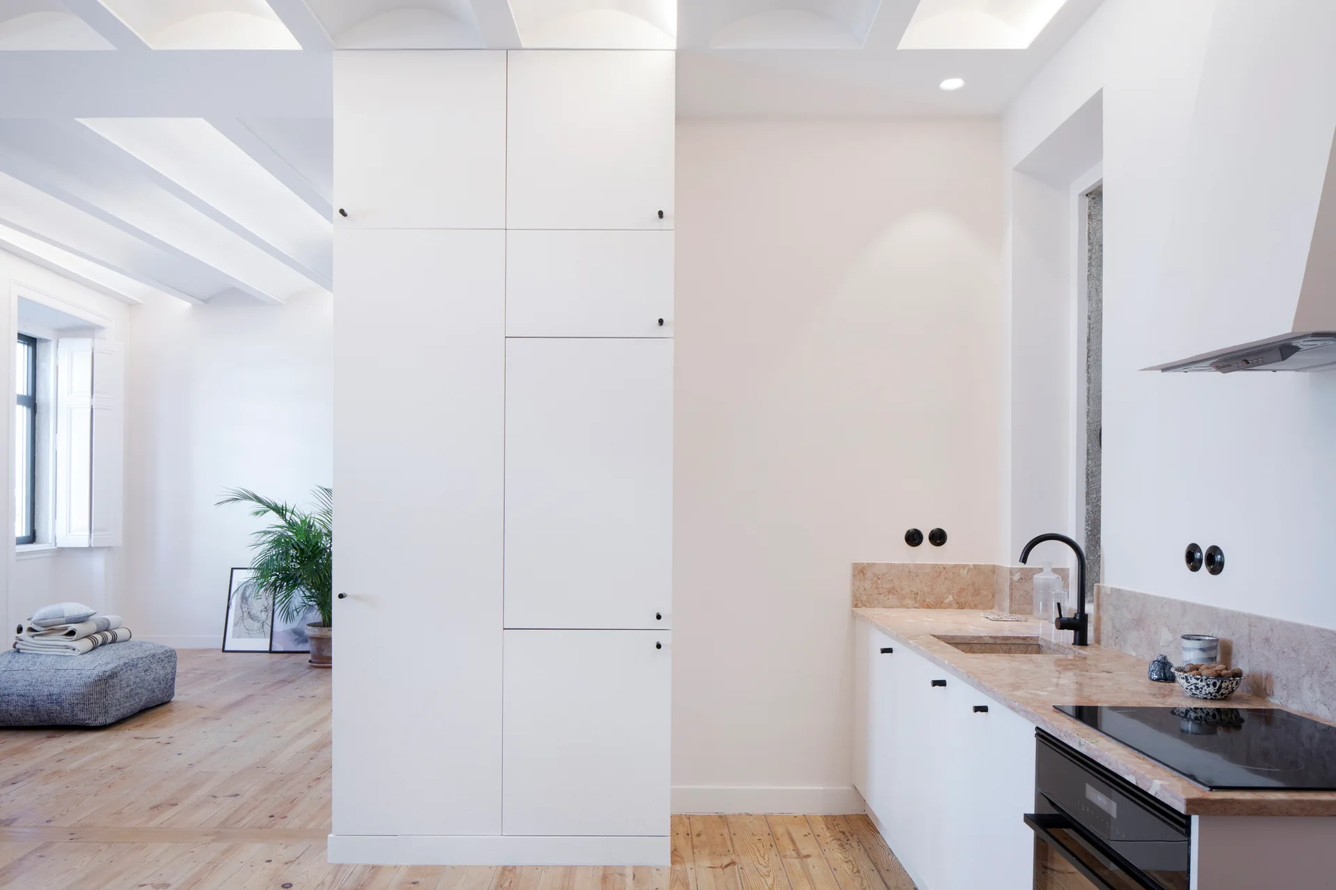 A minimalist kitchen featuring tall white pantry cabinets, a beige stone countertop, and light wood floors opening to a living room.