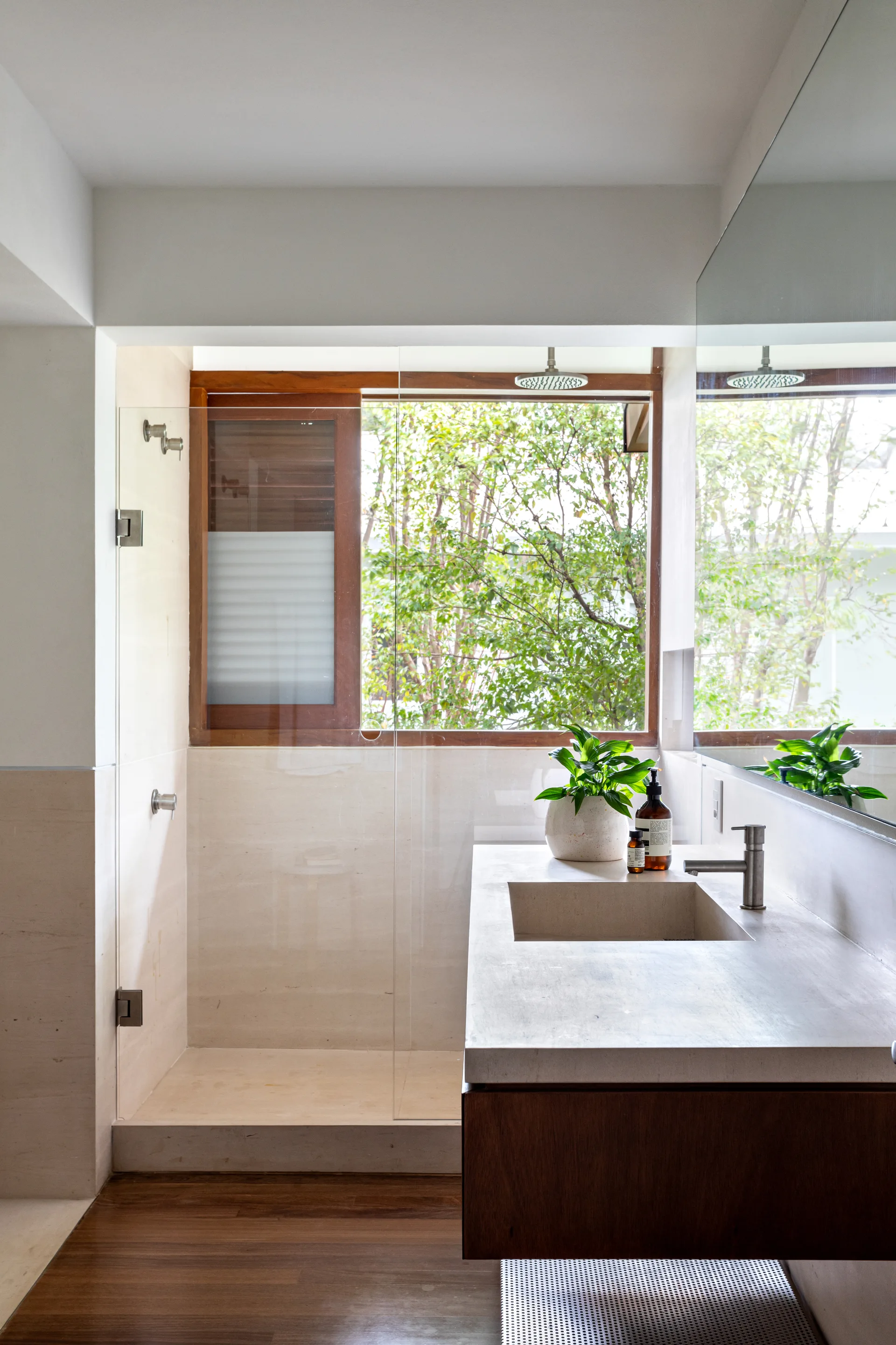 A modern bathroom features a stone-topped wood vanity next to a glass-enclosed shower with a large wooden-framed window overlooking trees.