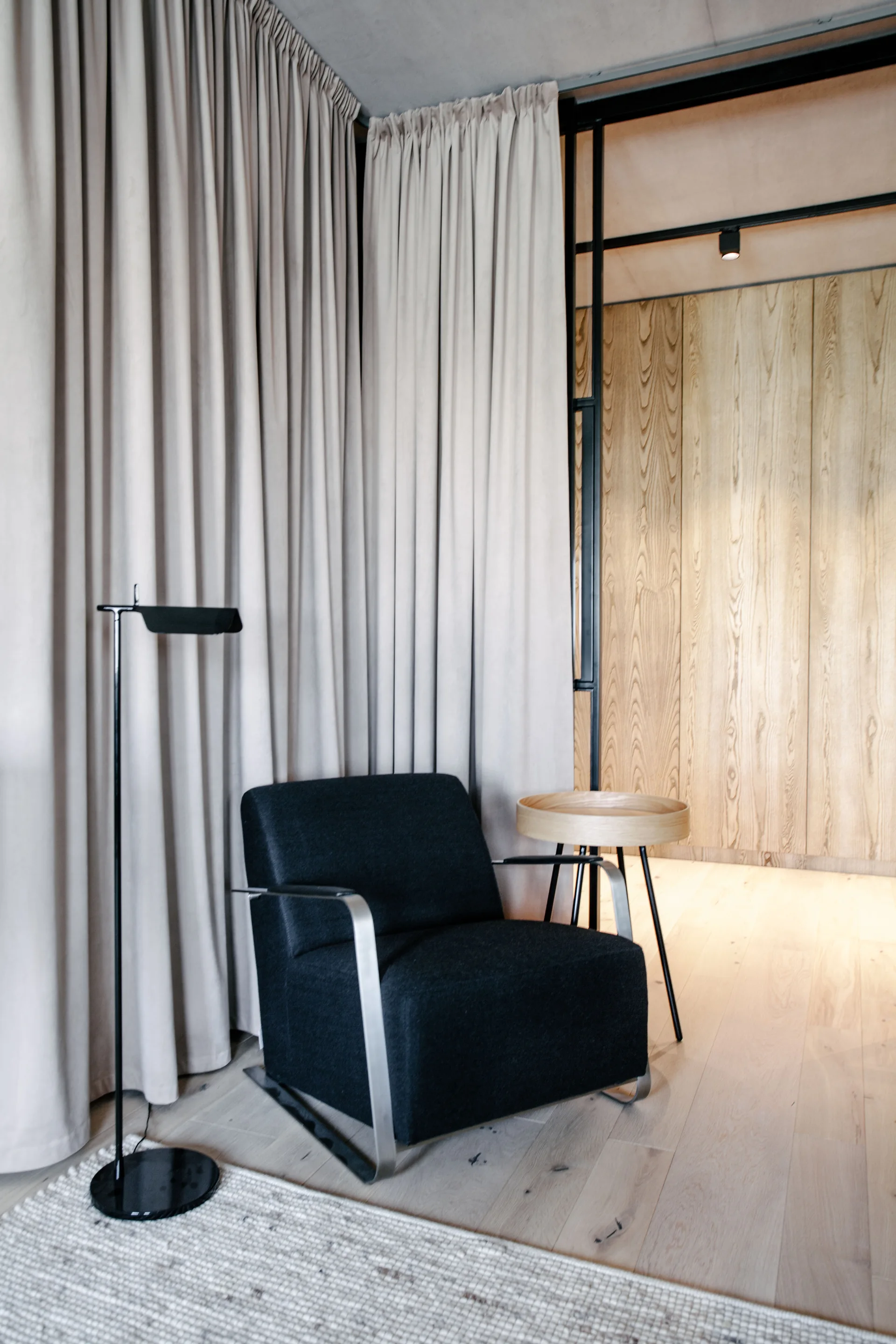 A dark upholstered armchair sits on a light woven rug next to a wooden side table, set against beige floor-to-ceiling curtains and a wood paneled wall.