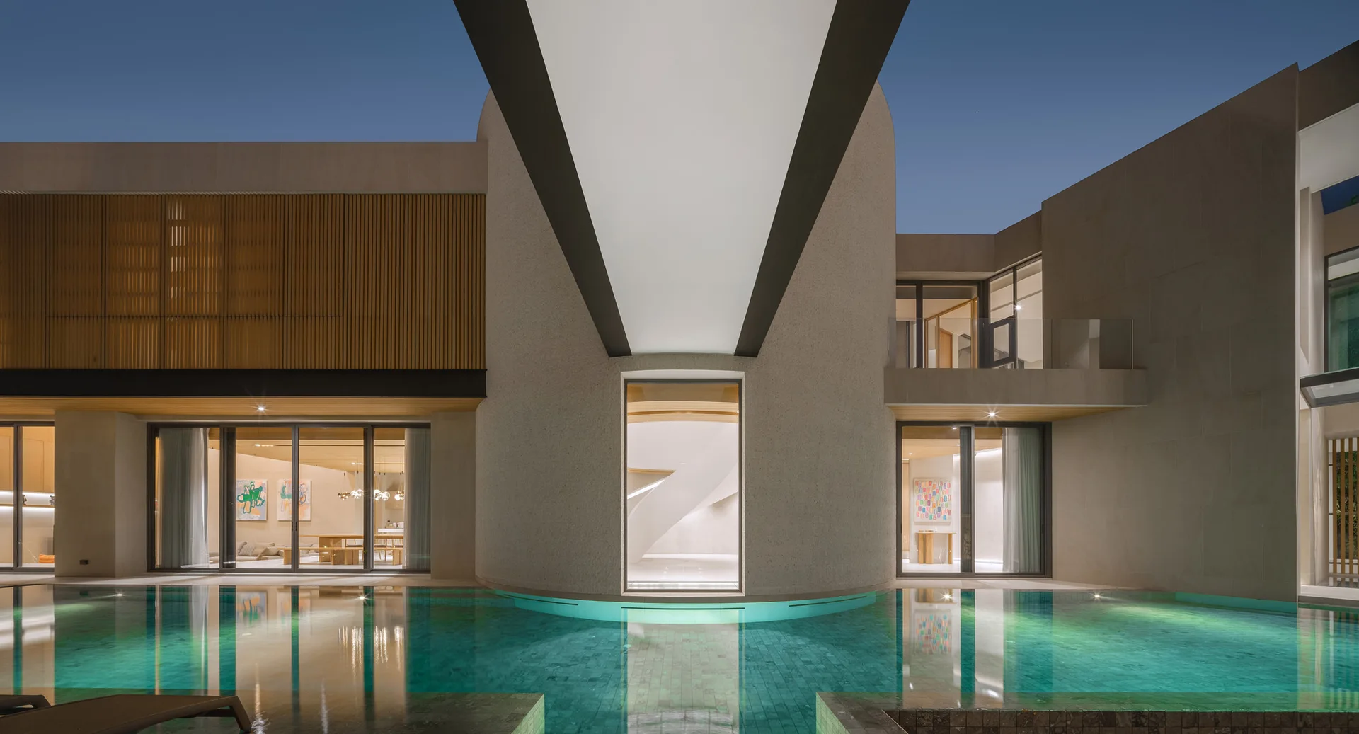 An illuminated swimming pool reflects a modern beige stone home with a central curved wall and large glass doors at dusk.