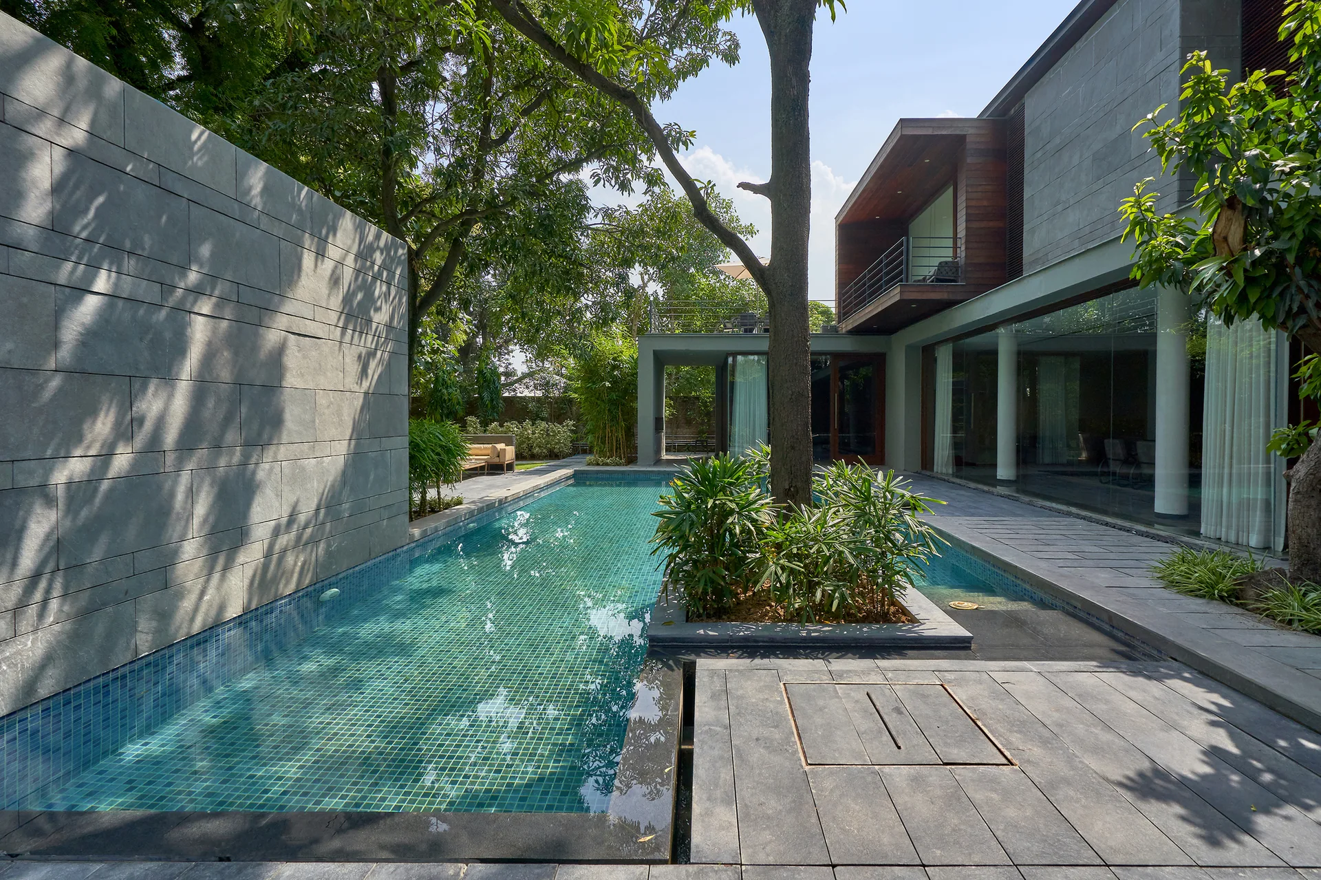 A rectangular backyard swimming pool sits between a tall gray stone wall and a modern glass-walled house with an integrated tree planter.