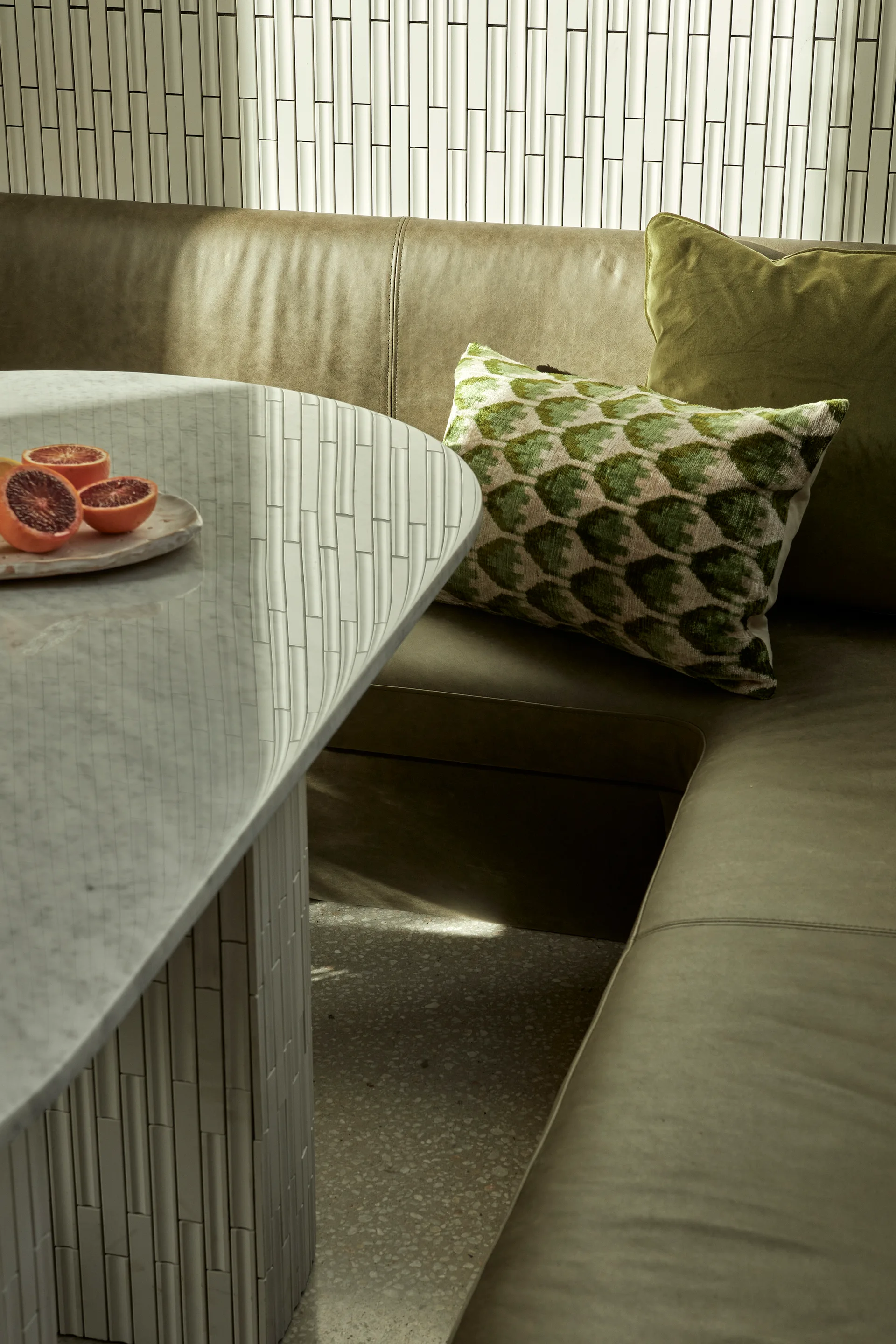 An olive green leather banquette sits next to an oval marble dining table with a tiled cylindrical base against a vertical tiled wall.