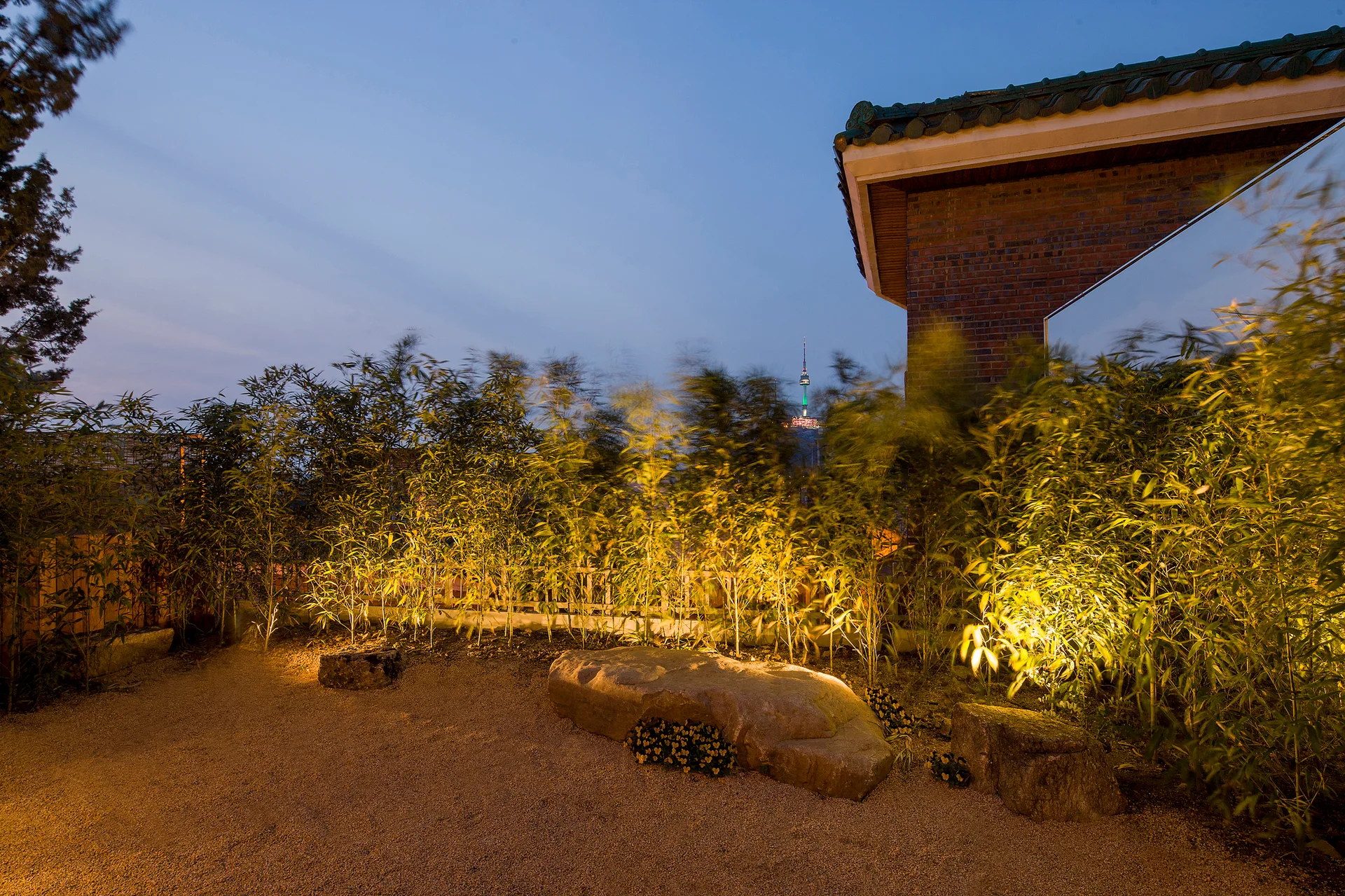 A courtyard garden at twilight featuring a border of illuminated bamboo, large natural stone boulders, gravel ground, and a brick building corner.