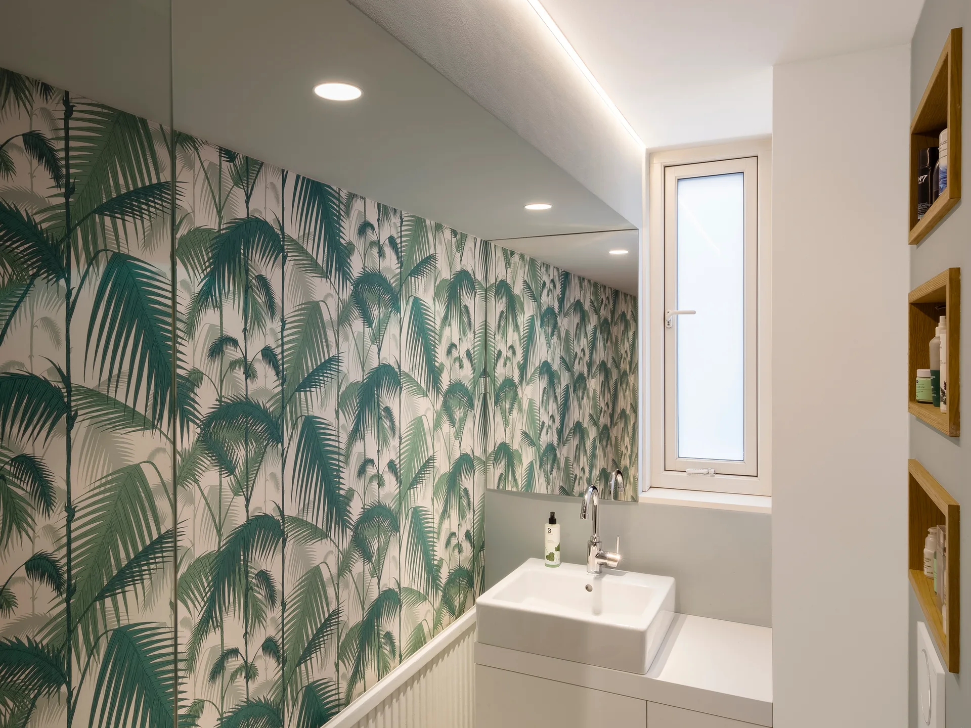 A small modern bathroom featuring green palm leaf wallpaper, a white vanity with a square basin, a large mirror, and recessed wooden wall shelves.
