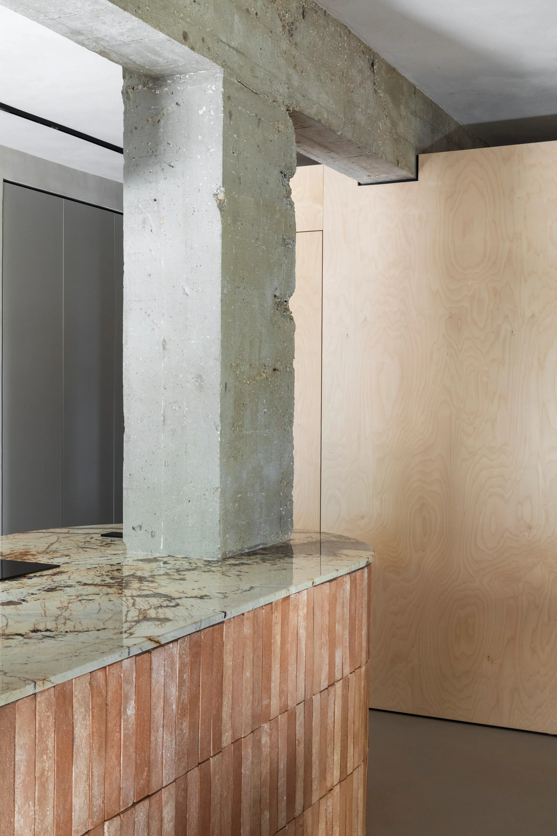 A curved kitchen island with a veined stone countertop and terracotta tile base stands beside a raw structural concrete column and plywood wall.
