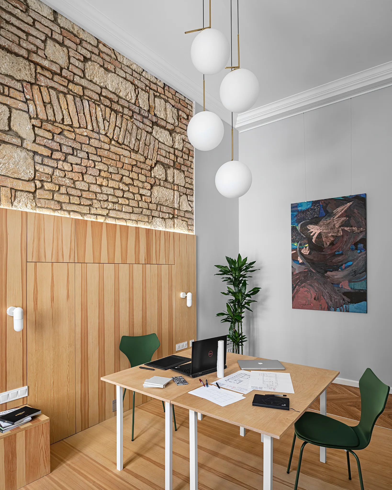 A modern home office features a wooden desk with green chairs, a wall with exposed brick and wood paneling, and globe pendant lights.