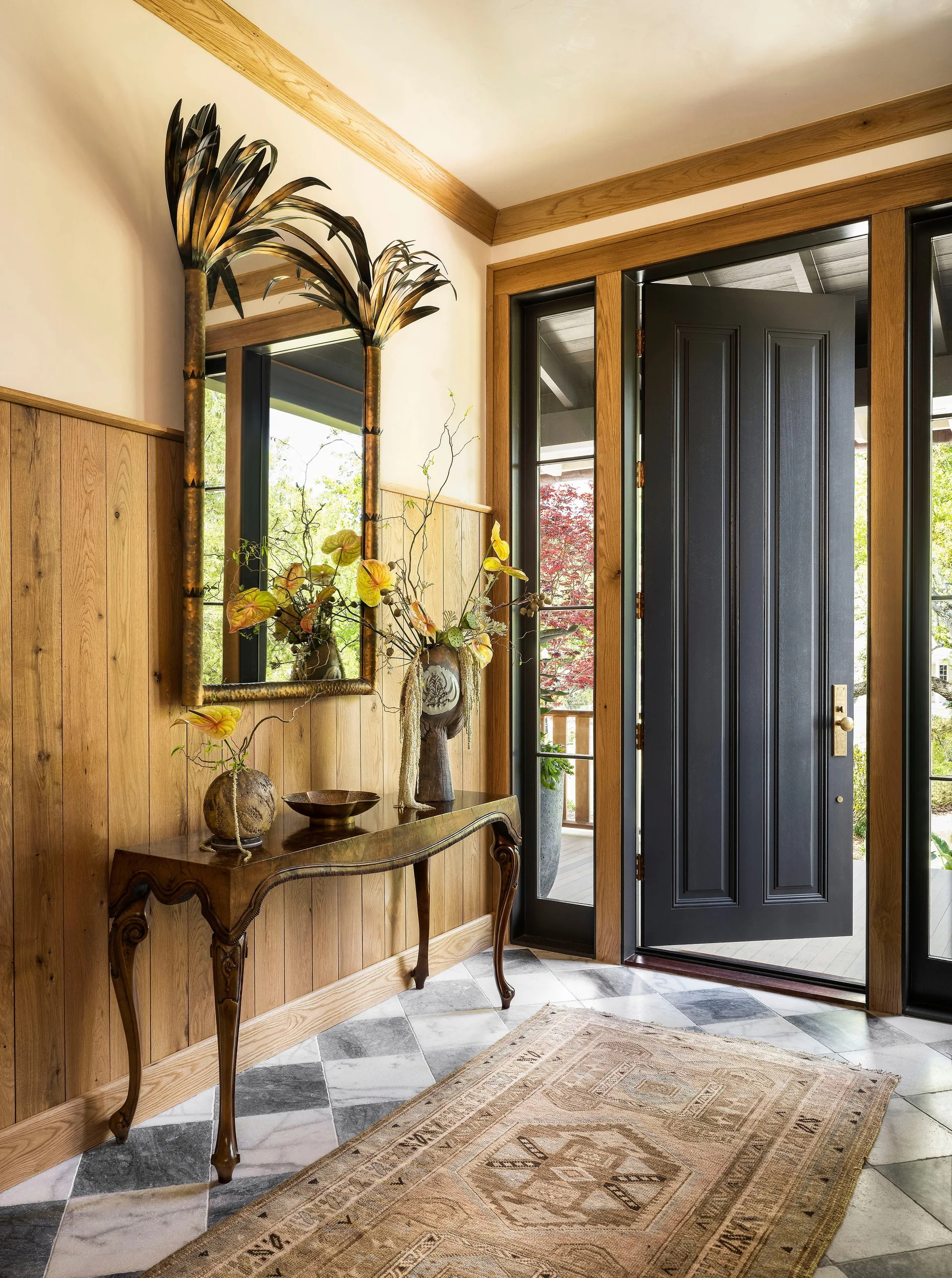 An entryway featuring vertical wood wall paneling, a black front door, a carved wood console table, and checkered marble floor tiles.