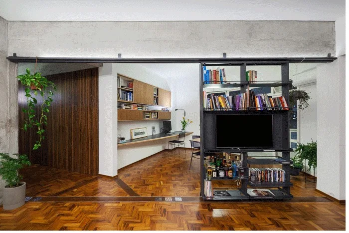 An open home office features a sliding black metal shelving unit, a long wood floating desk, and warm herringbone wood floors.