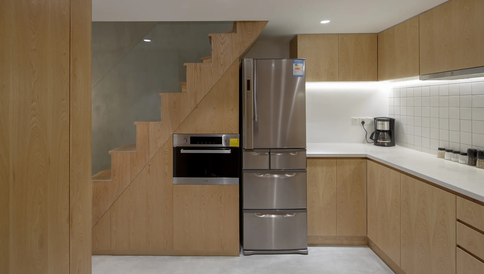 A modern kitchen with light wood cabinets, stainless steel refrigerator and built-in oven under a wood staircase, and white square tiled backsplash.