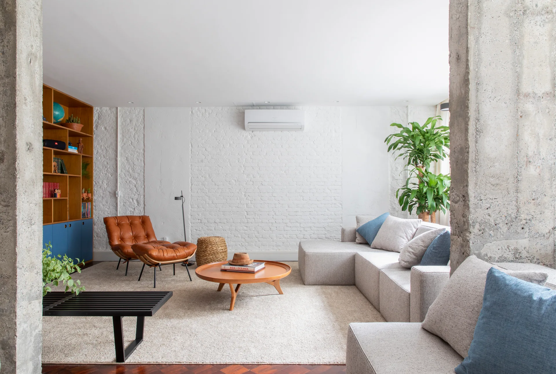 A light gray sectional sofa and a tan leather lounge chair sit on a beige rug in a living room with a white painted brick wall and concrete columns.