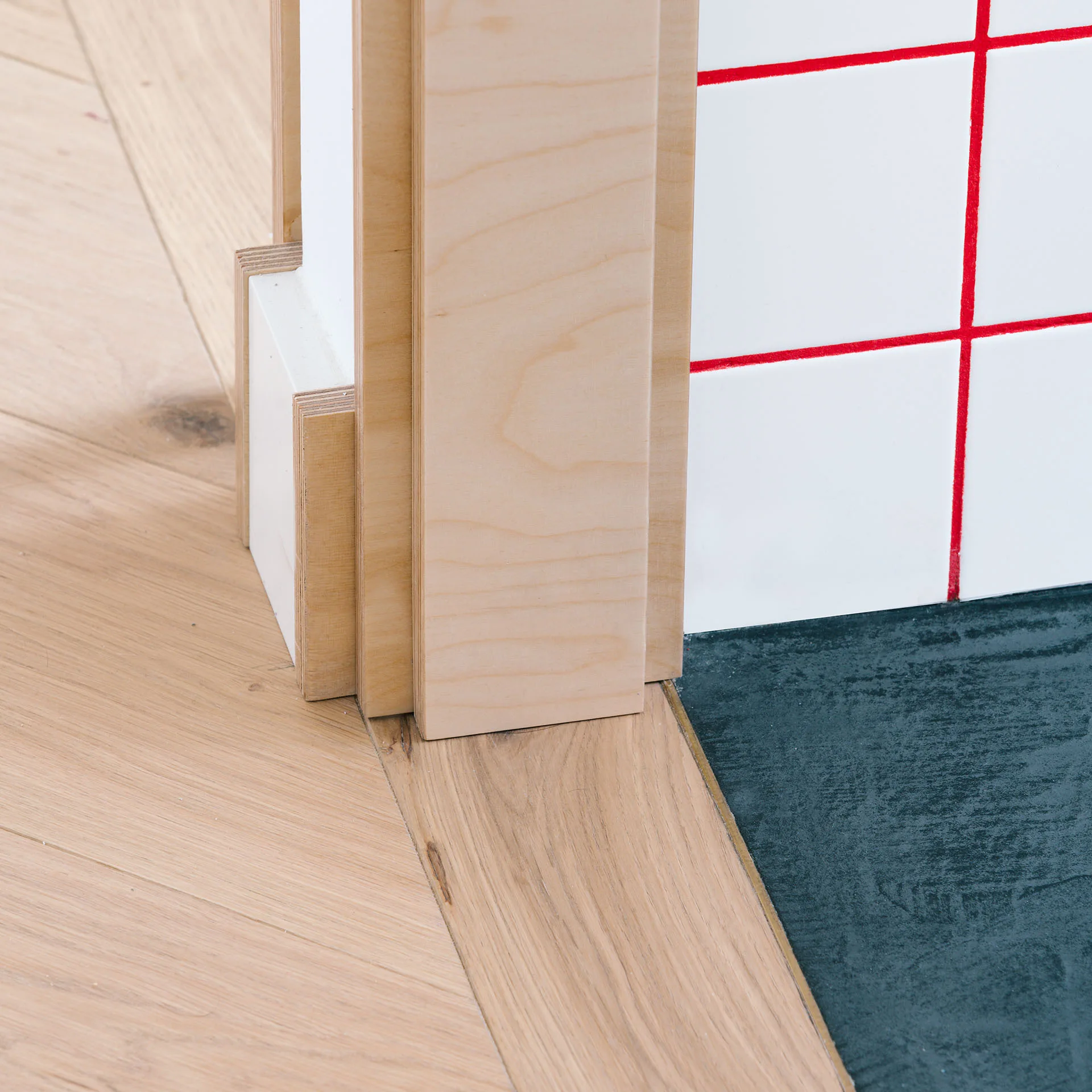 A close-up of a wall corner featuring layered plywood trim, white square tiles with bright red grout, herringbone wood floor, and dark slate flooring.