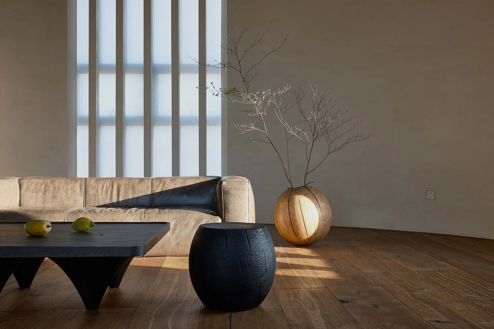 A minimalist living room features a tan leather sofa, a dark wood coffee table, a black barrel stool, and a spherical wood vase with dry branches.