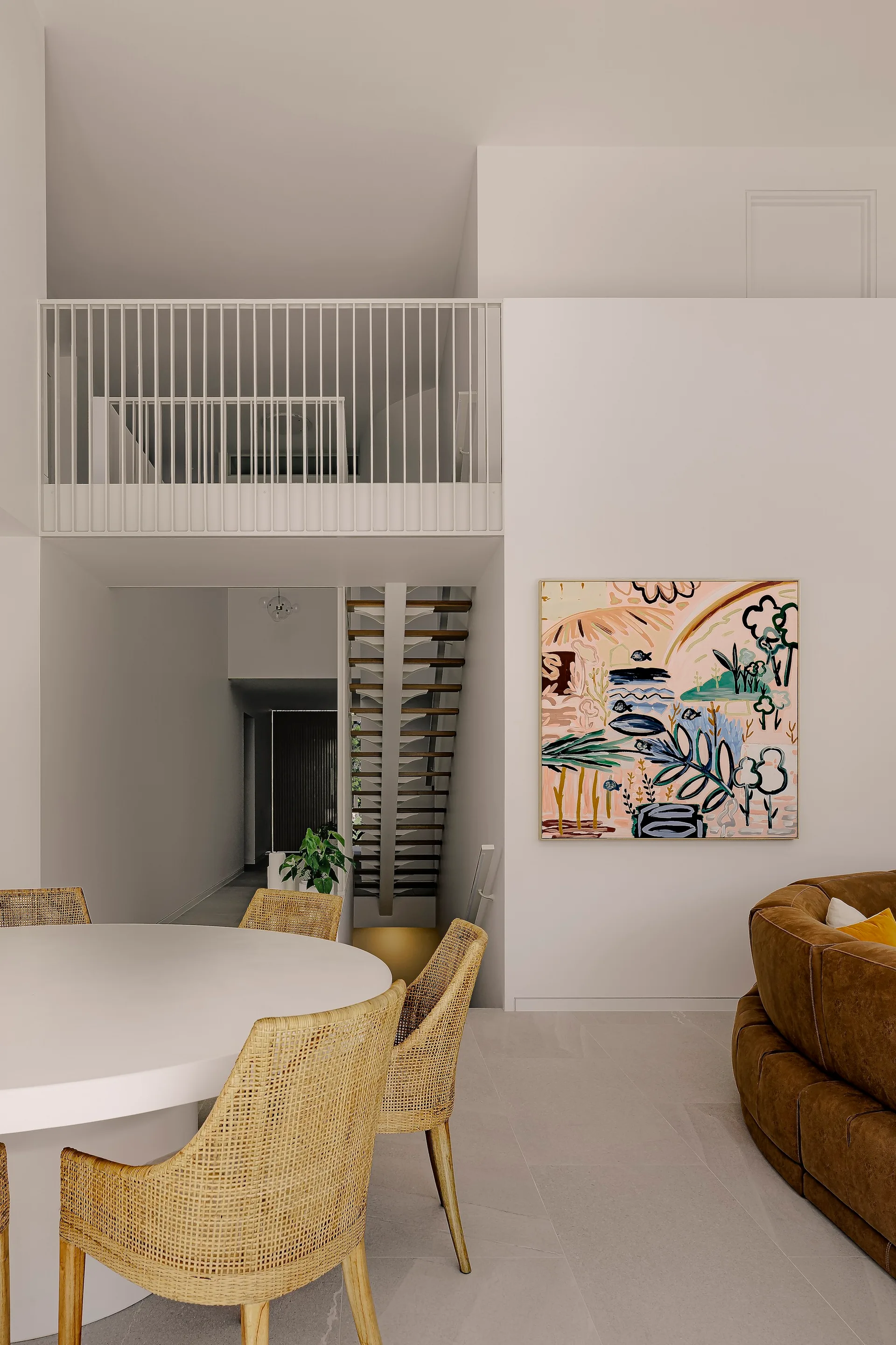 A modern open-concept space featuring a white dining table with woven rattan chairs, a curved brown leather sofa, and an open-riser staircase.