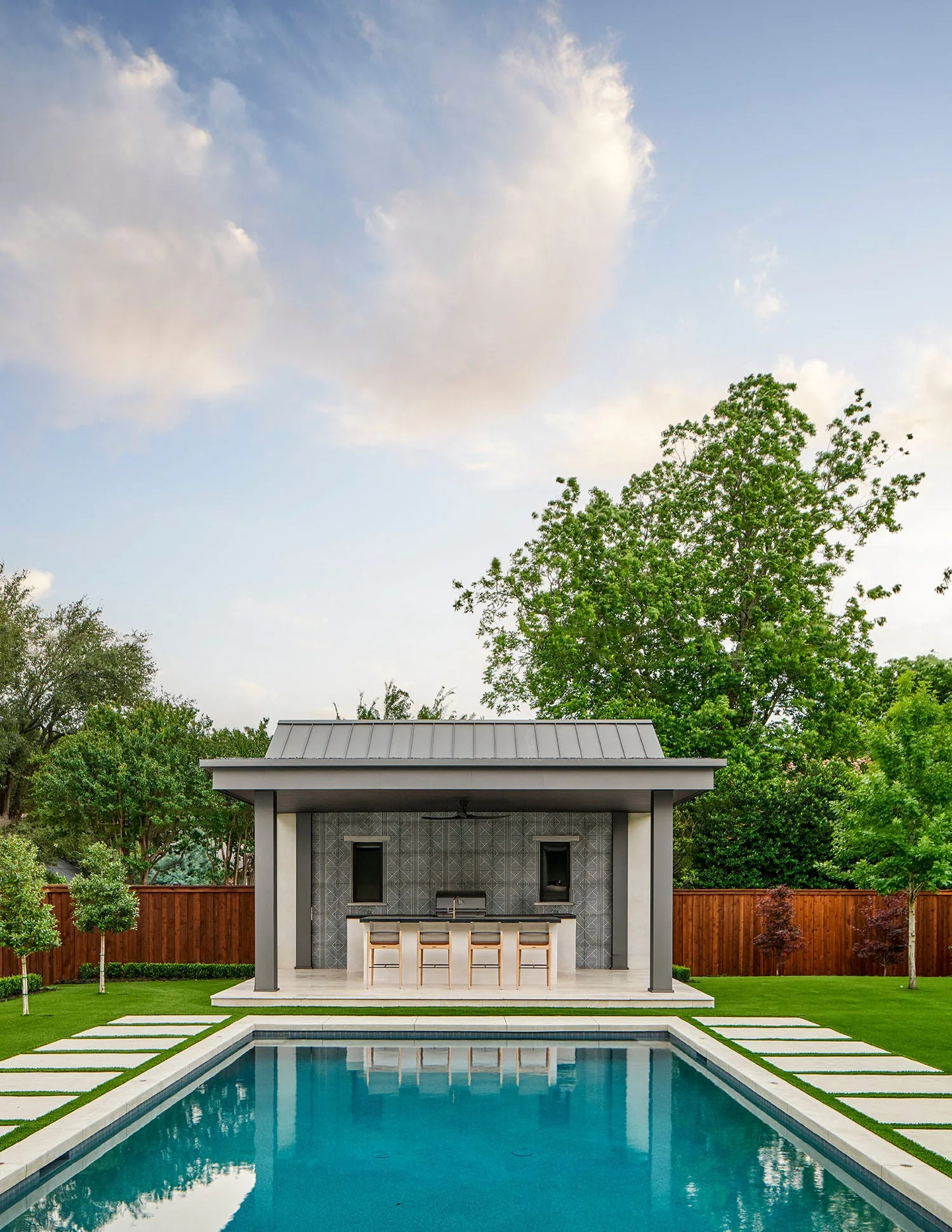 A covered modern outdoor kitchen with a geometric tile wall and bar stools sits behind a rectangular blue swimming pool in a lush backyard.