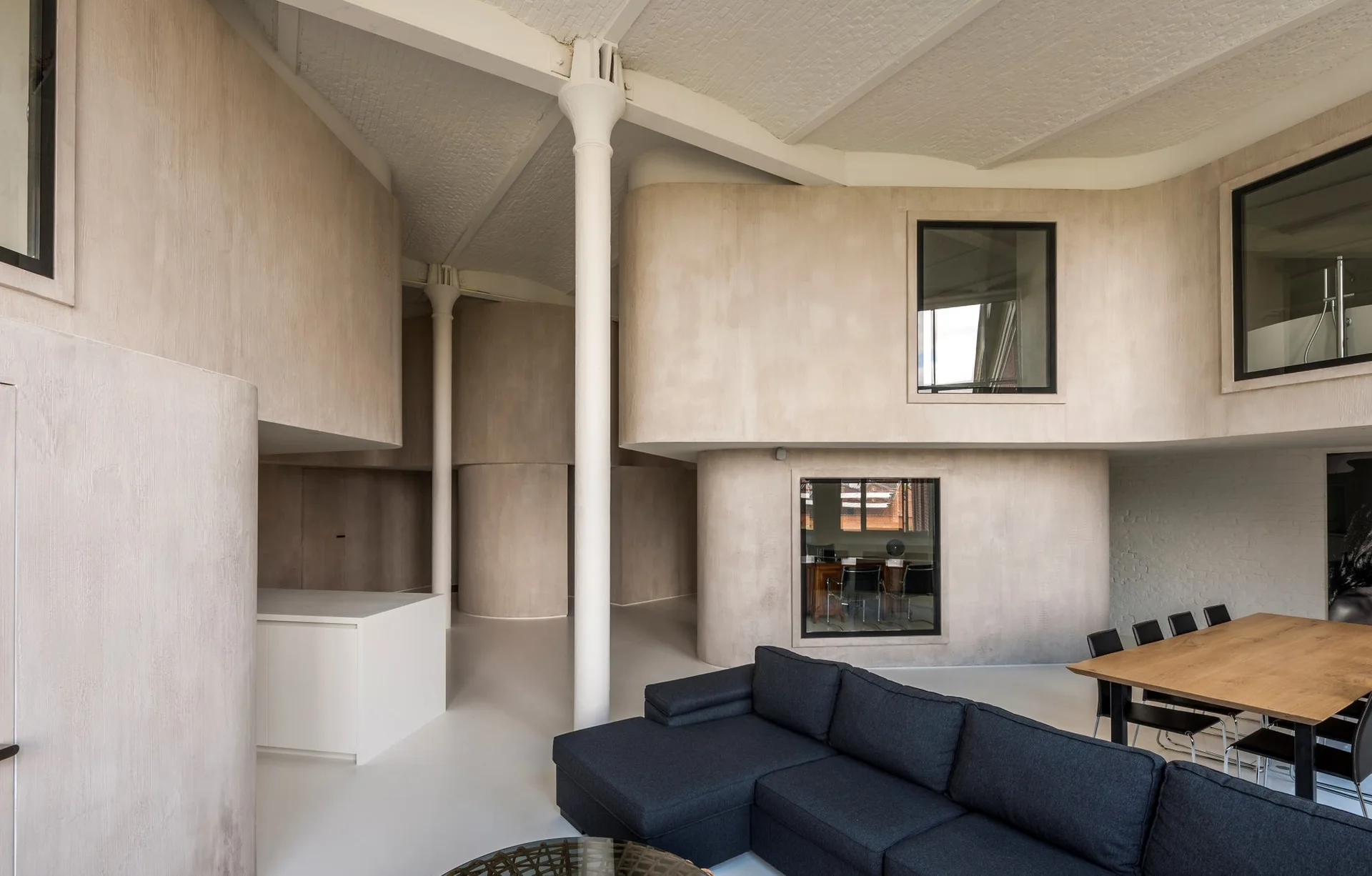 A spacious modern living area featuring a dark gray sectional sofa, a wooden dining table, white structural columns, and curved beige plaster walls.