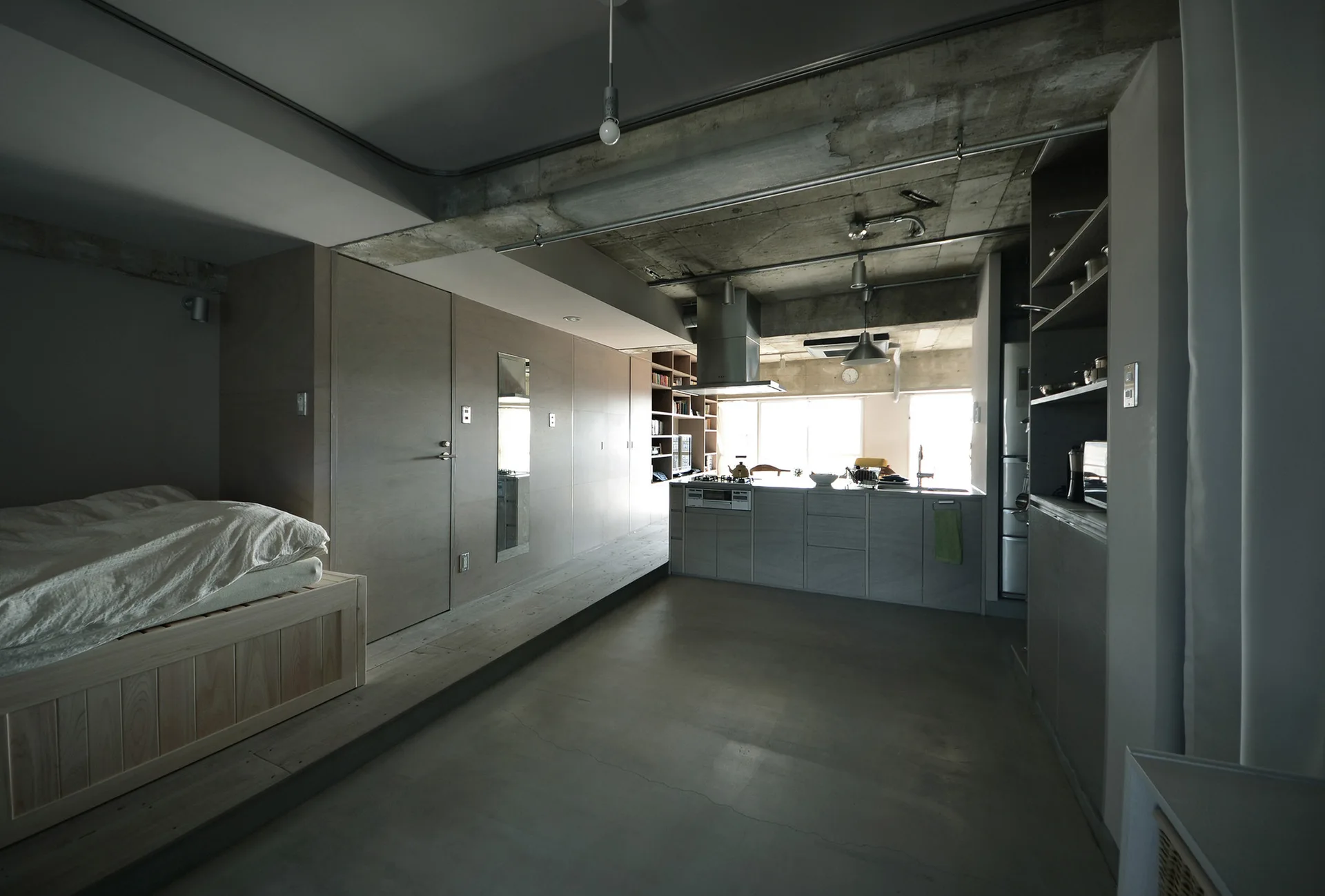 An open-plan loft featuring an exposed raw concrete ceiling, a minimalist kitchen island, and a bed on a raised wooden platform.