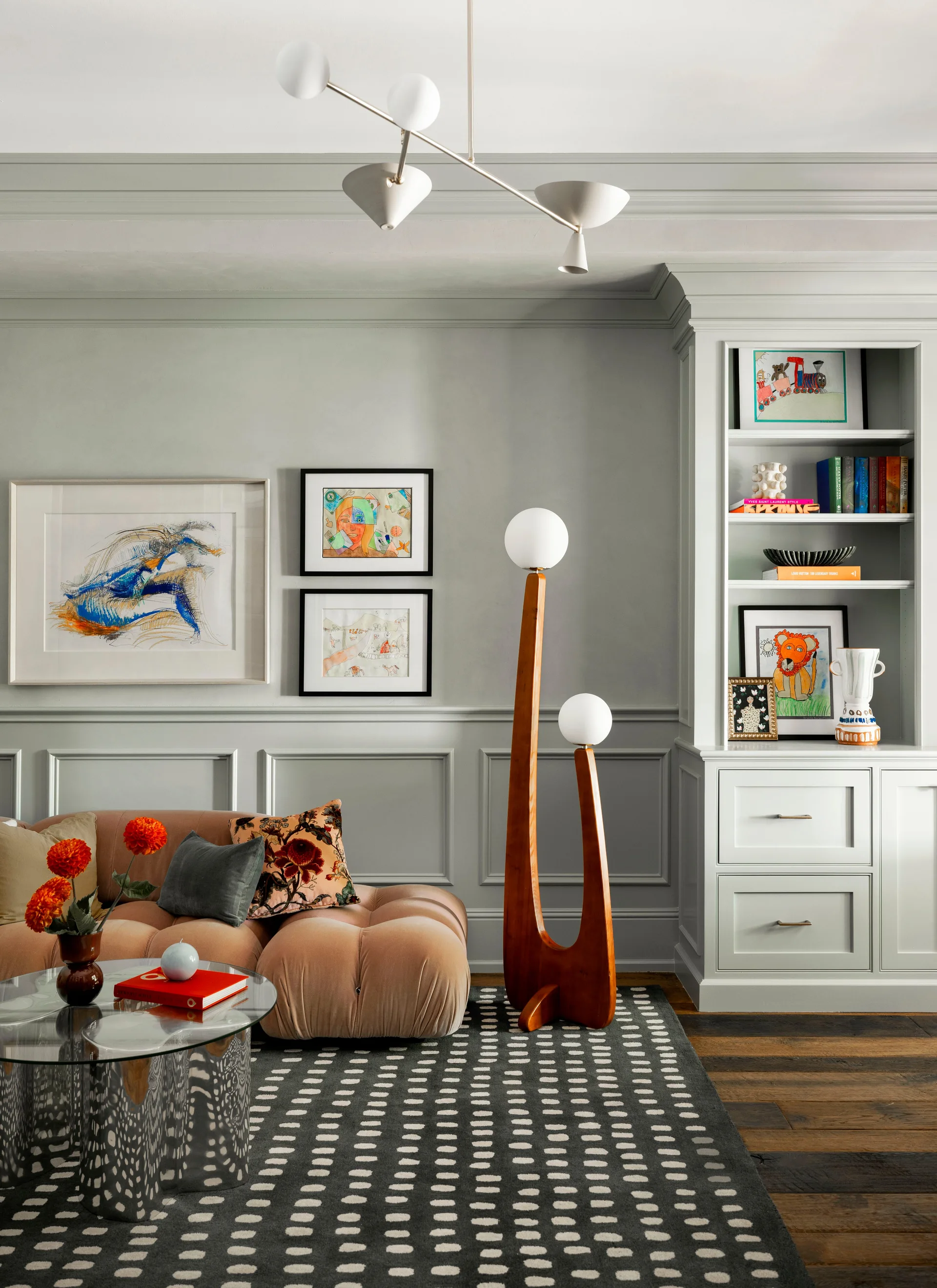 A living room featuring a pink tufted velvet sofa, a sculptural wood floor lamp, and white built-in shelving against gray paneled walls.