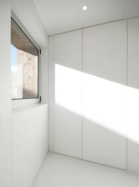 A bright minimalist interior corner features seamless white built-in cabinetry next to a window casting sharp natural light across the floor.