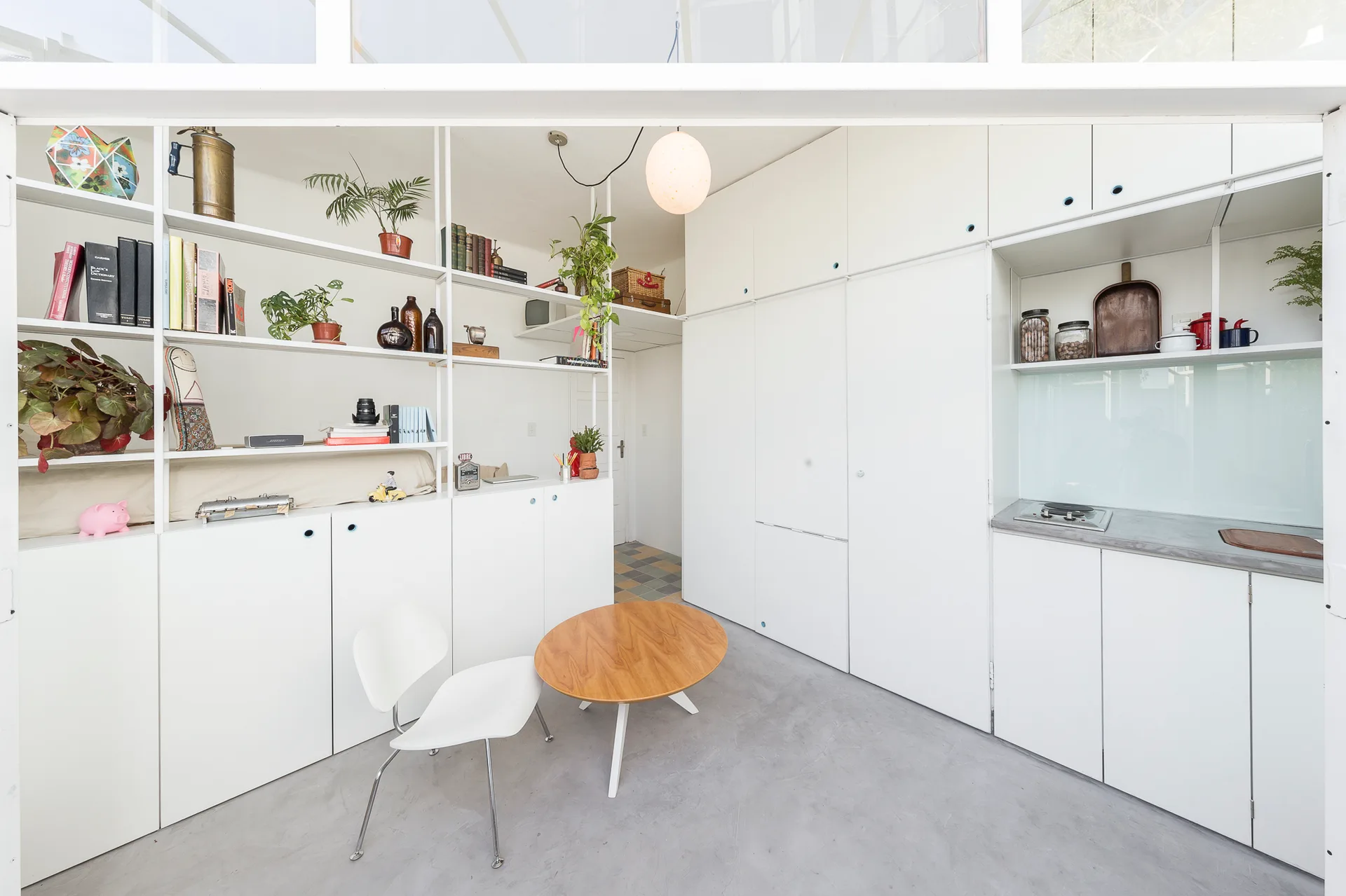 A minimalist white studio room featuring floor-to-ceiling cabinets, open metal shelving, and a small round wood table with two chairs.