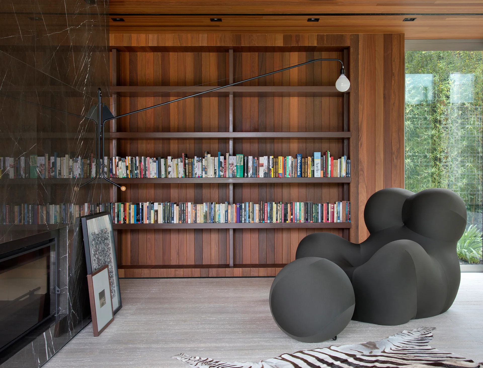 A modern home library featuring wood-paneled walls, built-in bookshelves filled with books, a dark gray sculptural armchair, and a black marble fireplace.
