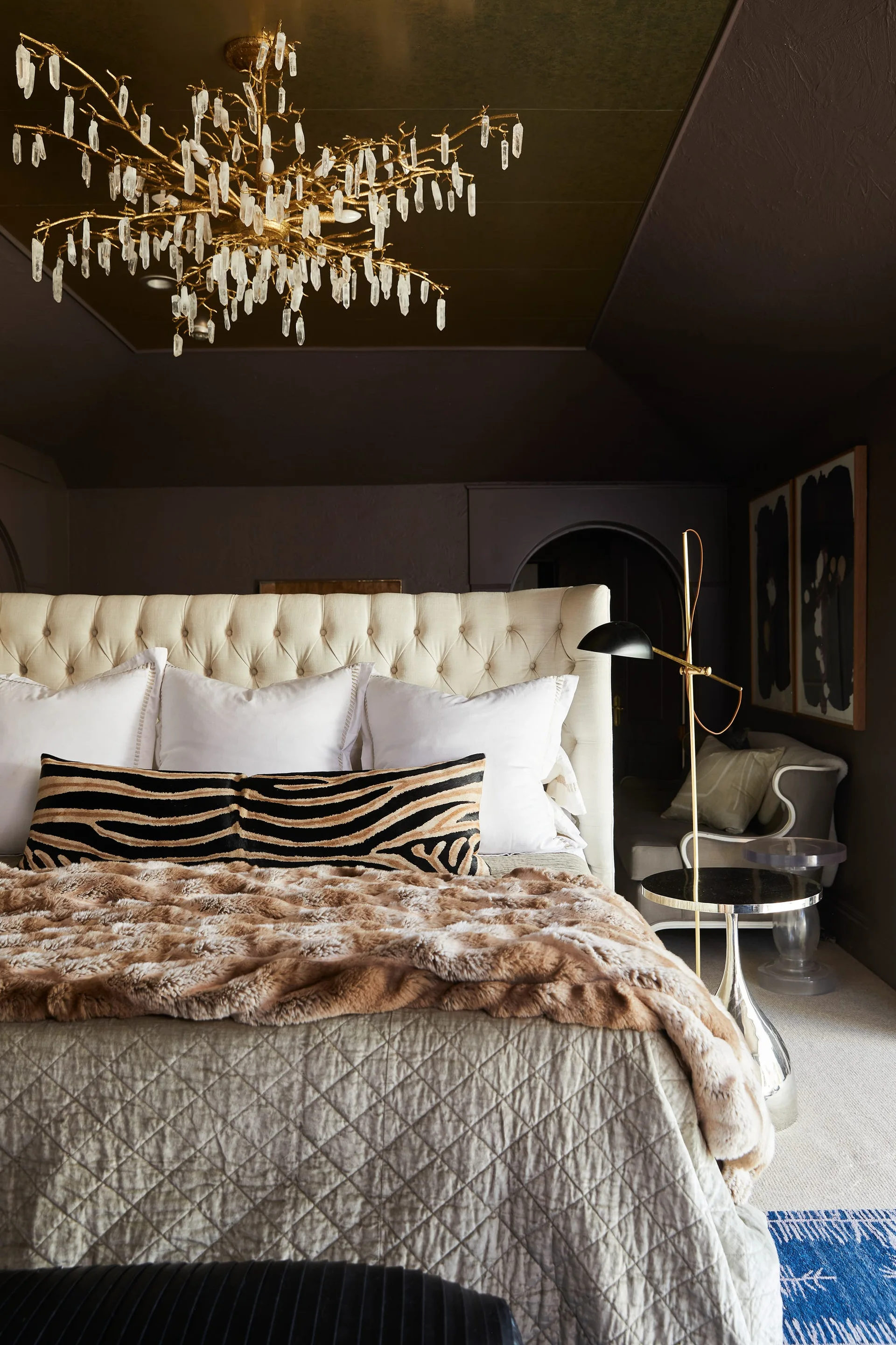 A dark bedroom features a cream button-tufted headboard, animal print bolster pillow, and an elaborate gold branch chandelier with hanging crystals.