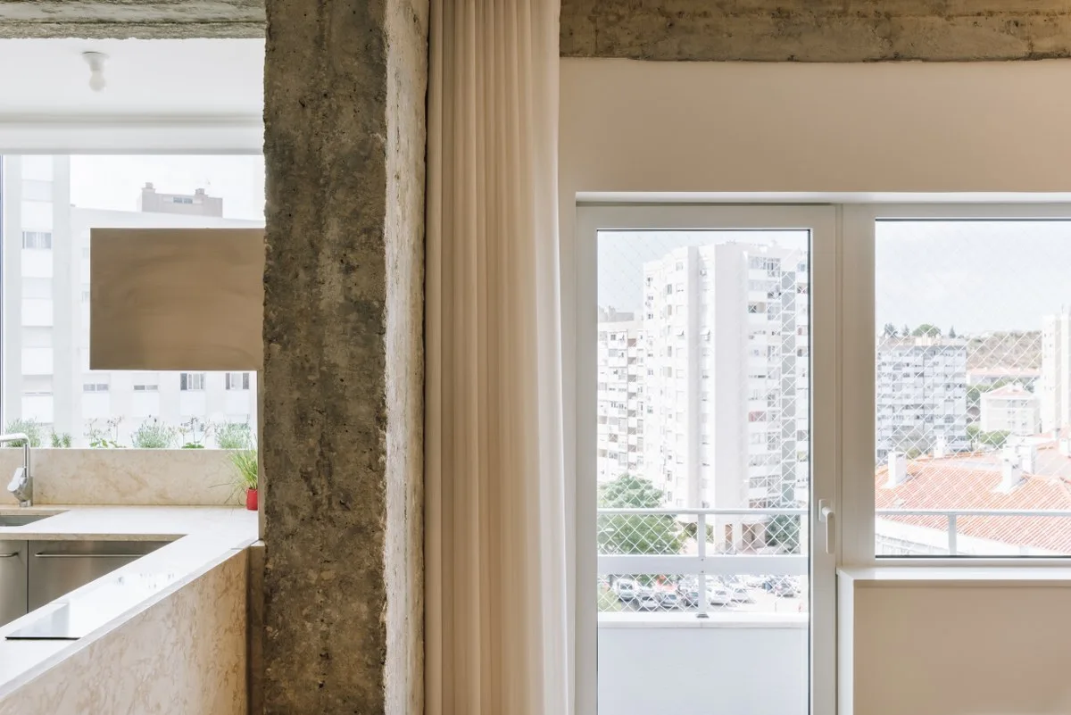 A bright kitchen features an exposed rough concrete column alongside a stone countertop and a white glass door opening to a balcony.