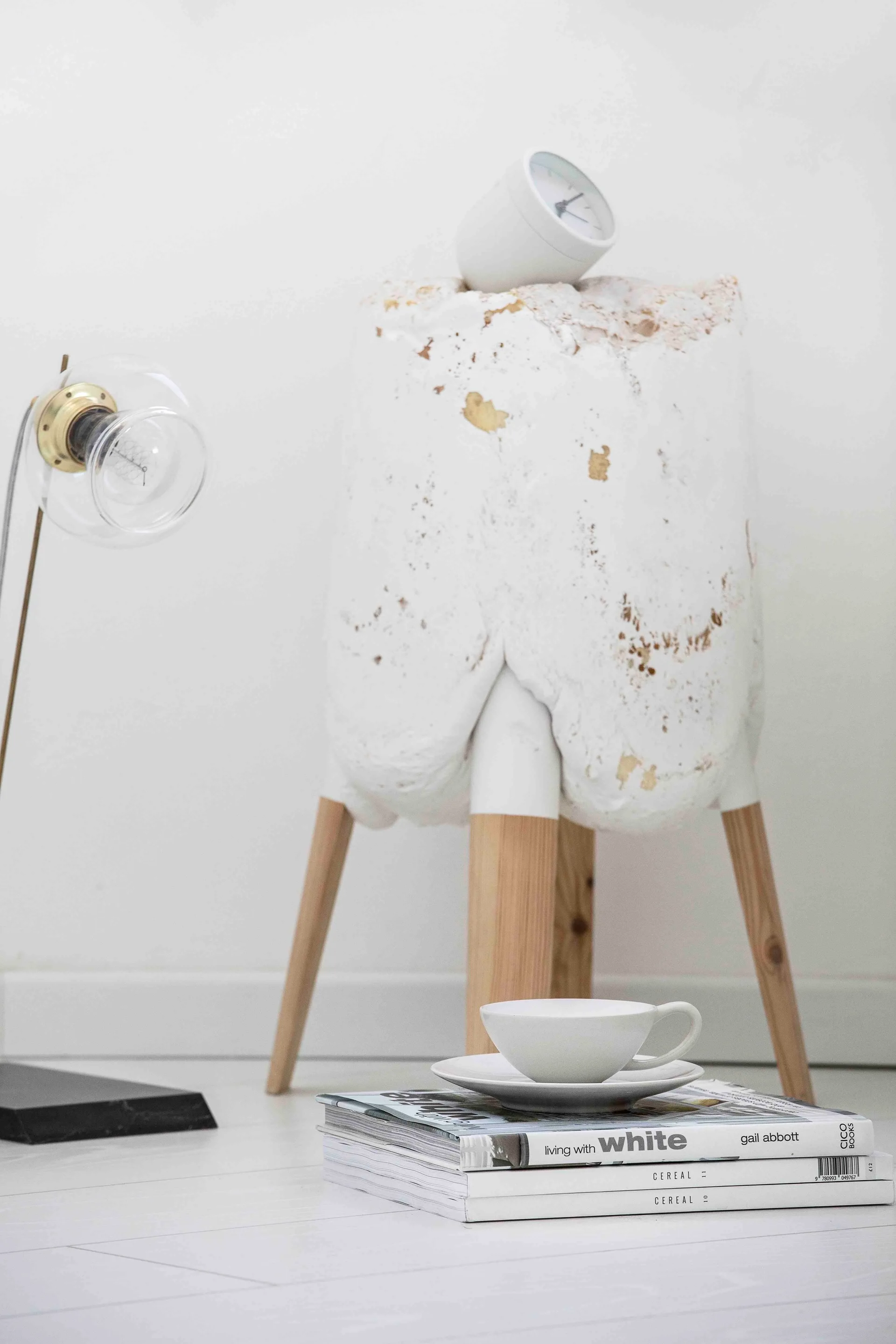 A sculptural white textured stool with wooden legs stands next to a glass table lamp and a stack of magazines topped with a ceramic teacup.