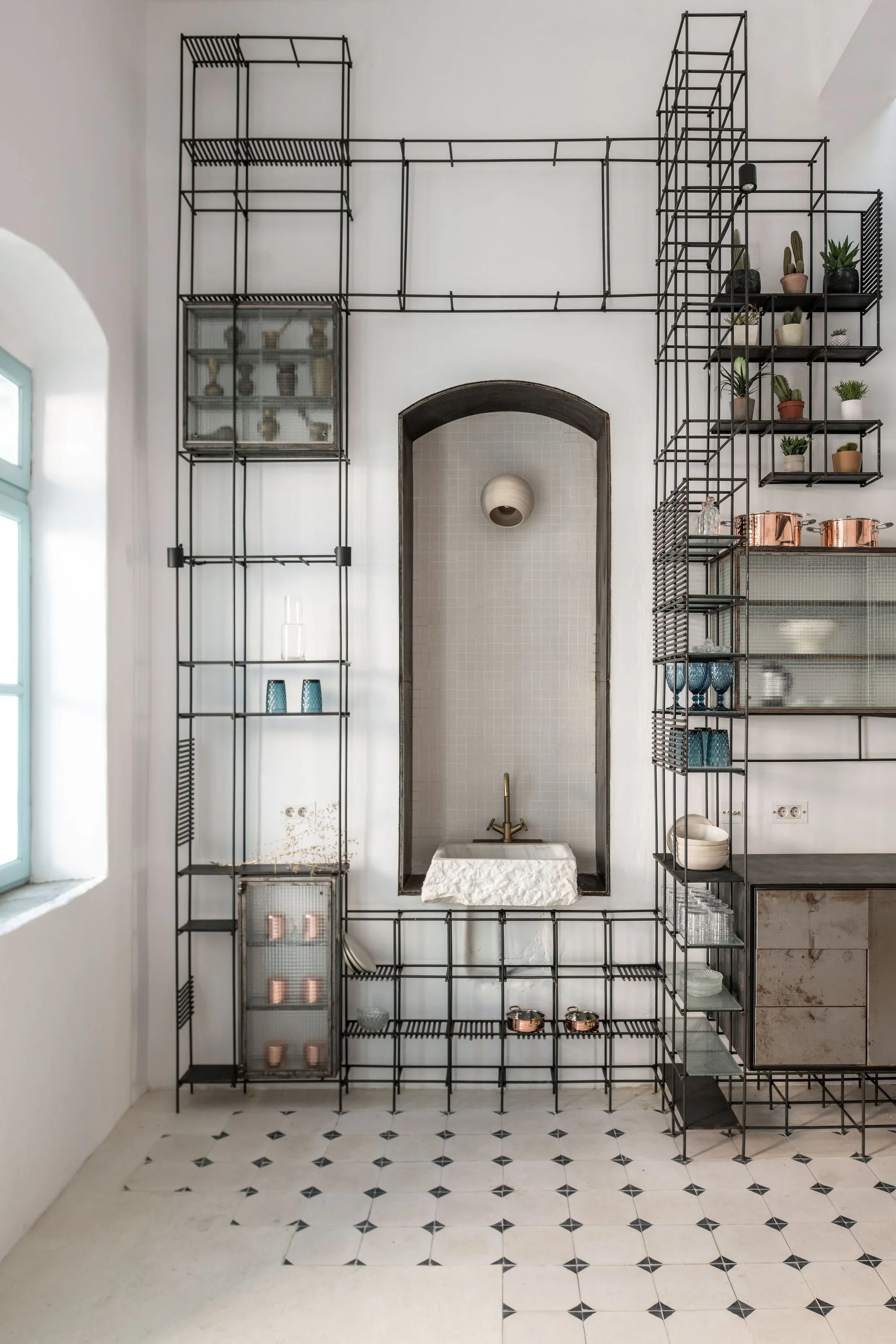 A raw stone sink sits in an arched tiled recess surrounded by tall black metal grid shelving on a patterned tile floor.