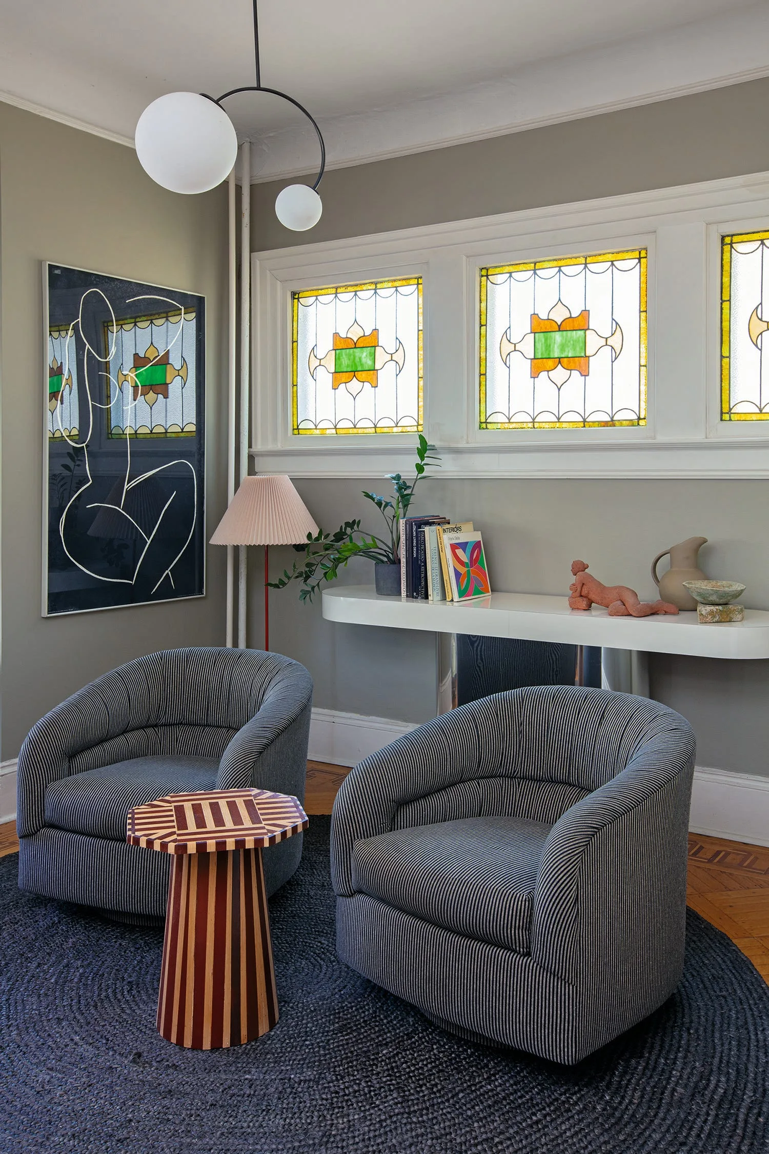 A sitting area features two gray pinstriped armchairs, a geometric wooden side table, a dark round rug, and stained glass windows on a gray wall.