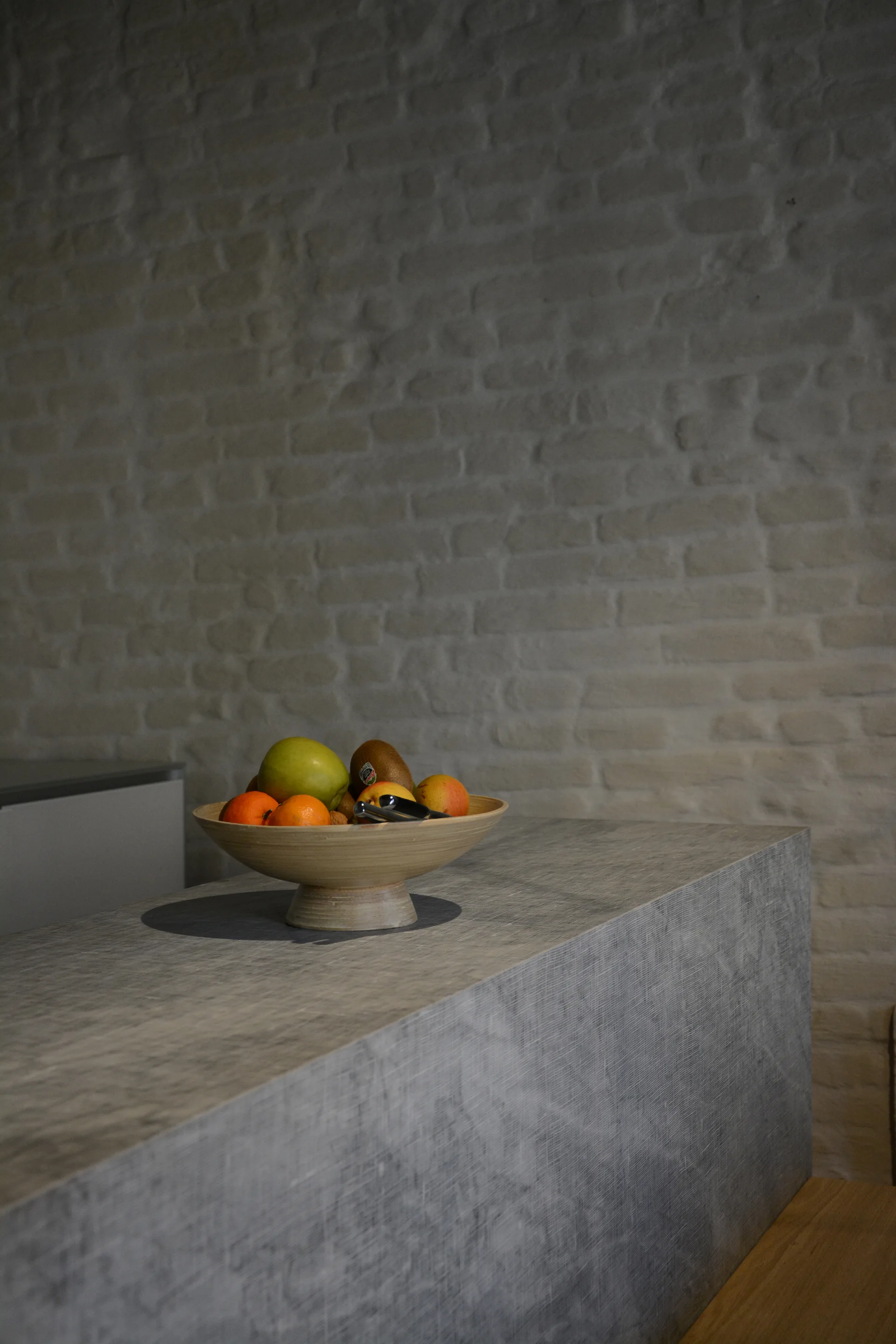 A pedestal fruit bowl rests on a textured gray kitchen island in front of a painted white brick wall.