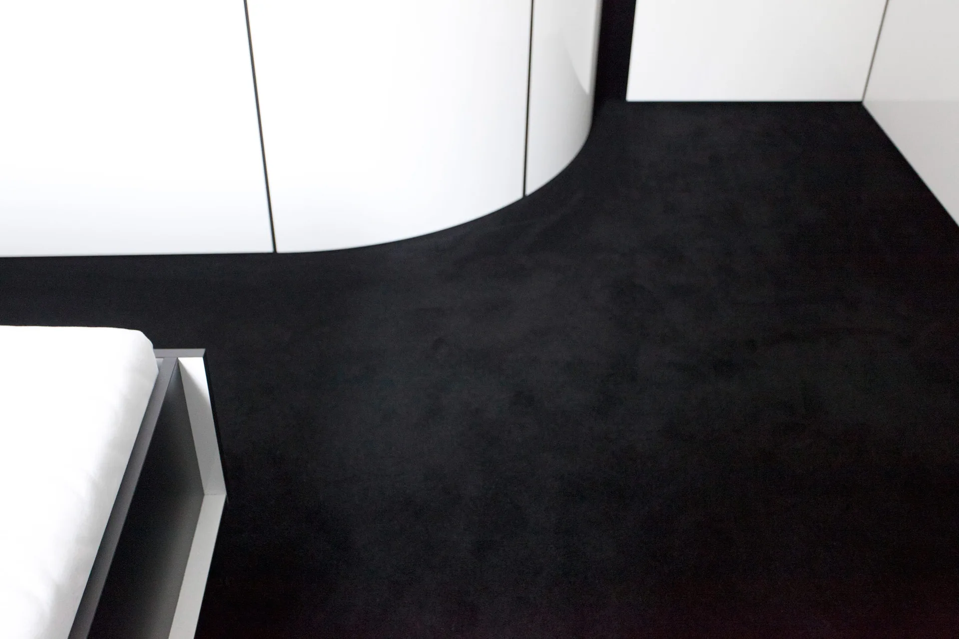 A high-contrast minimalist interior showcasing matte black carpeted flooring meeting a curved glossy white wall with thin vertical lines.