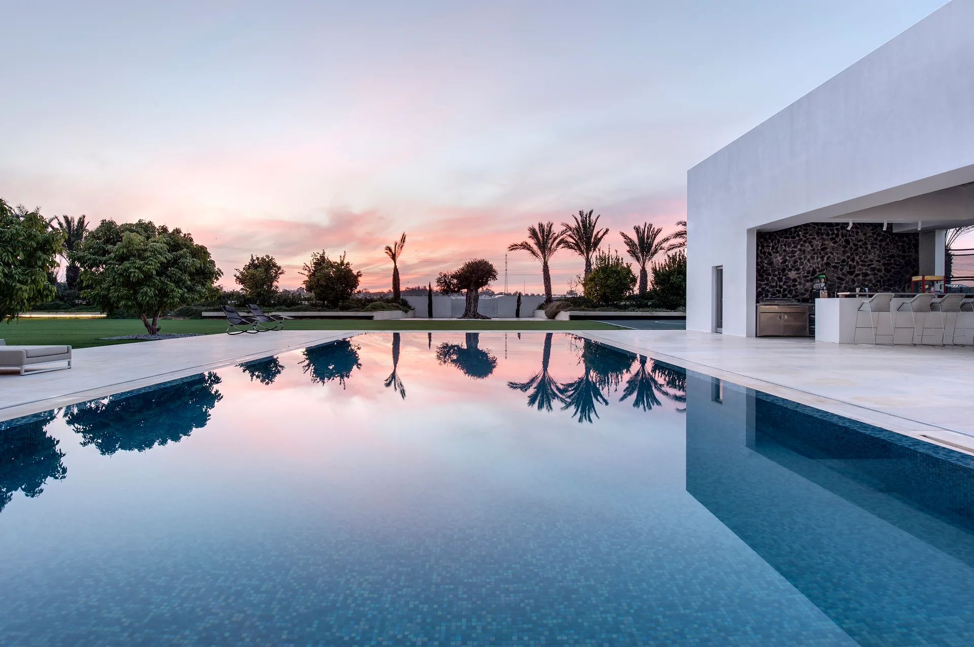 A rectangular pool reflects a pink sunset sky next to a modern white pavilion with an outdoor kitchen and bar stools.
