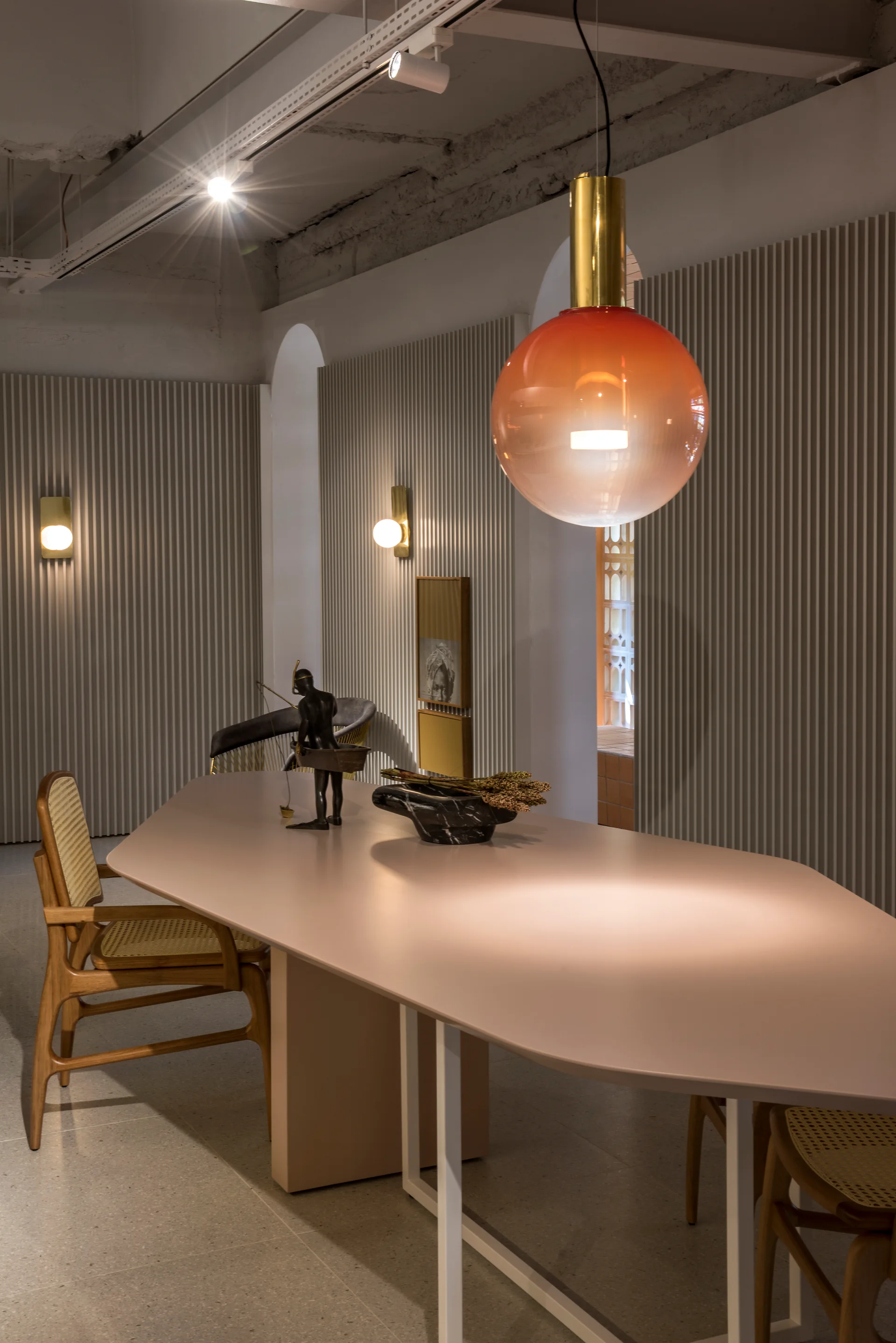 A modern dining area features a geometric pink table, cane chairs, and grey fluted wall paneling beneath a glowing orange glass pendant.