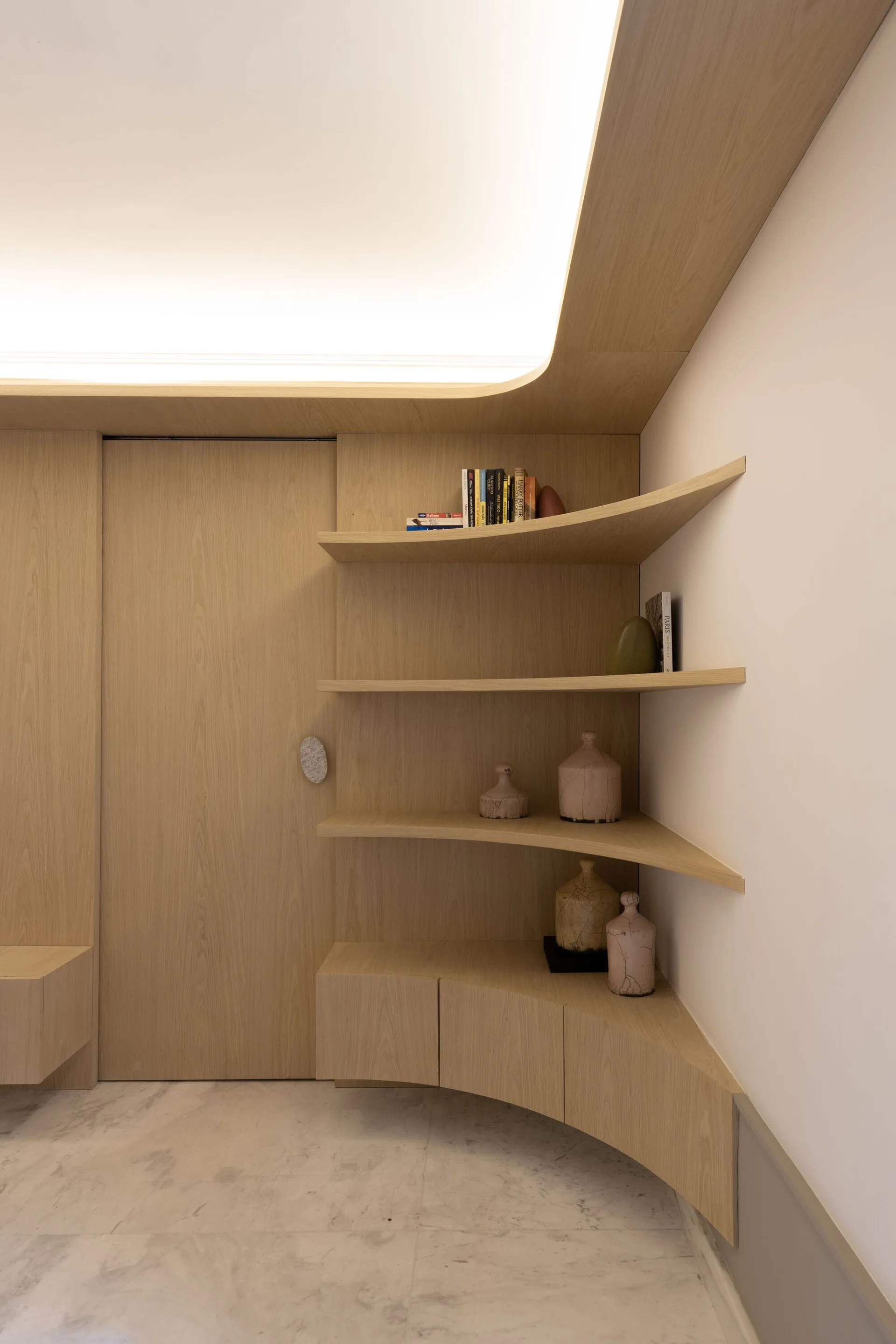 A minimalist curved wood storage unit features open shelves with ceramic vases, integrated lower cabinetry, and warm ceiling cove lighting.