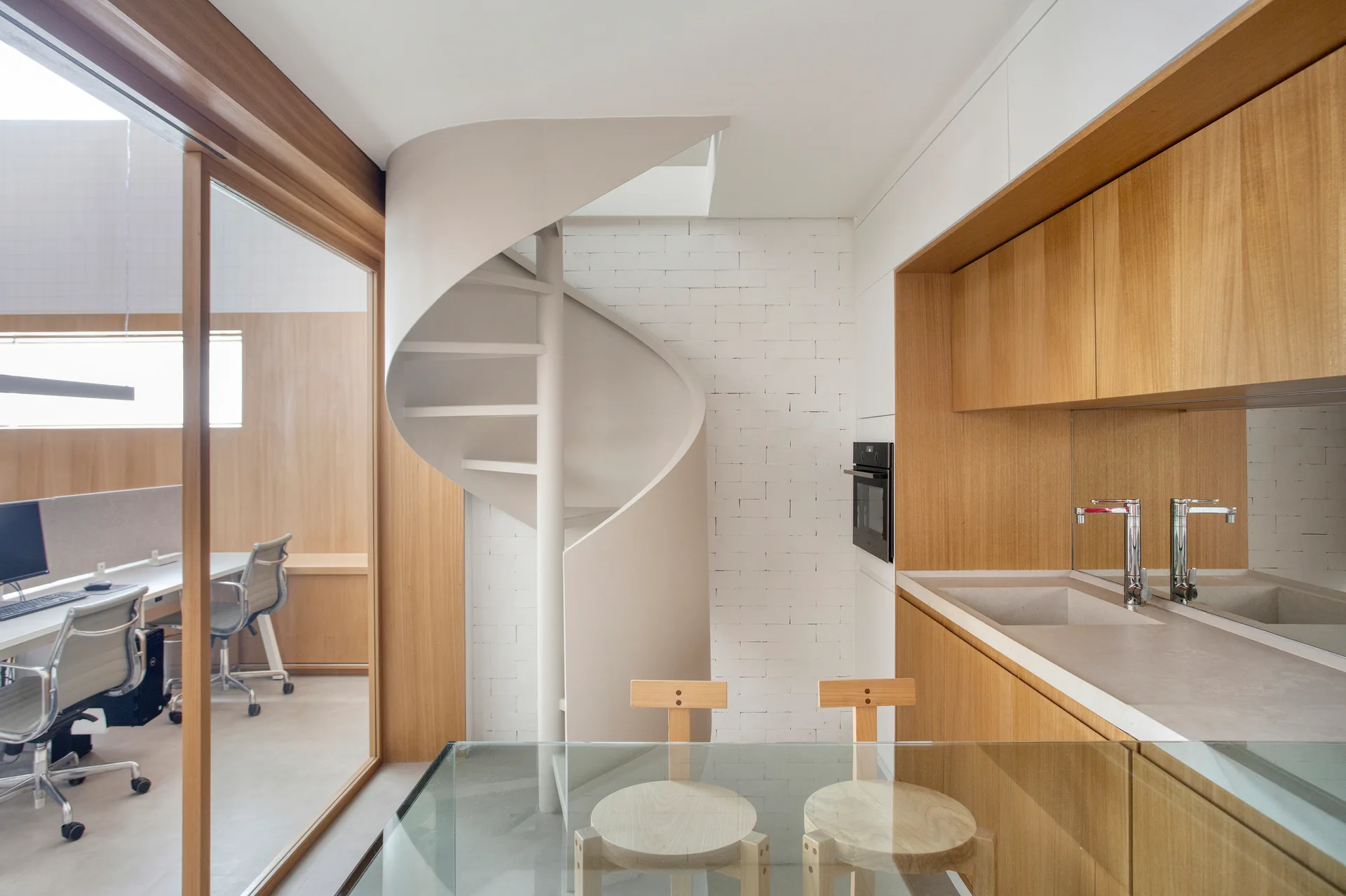 A modern kitchen featuring warm wood cabinetry, a white spiral staircase against a brick wall, and a glass-enclosed office space.